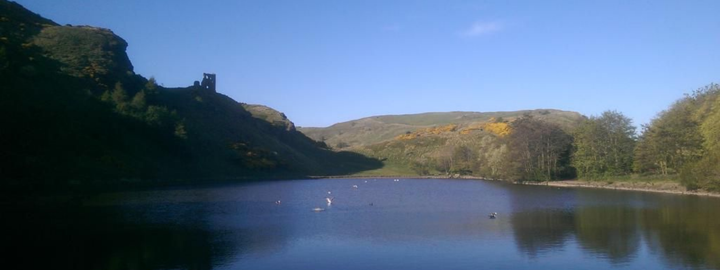 A peaceful lake surrounded by green hills with a ruin on top of a hill on the left side, under a clear blue sky.