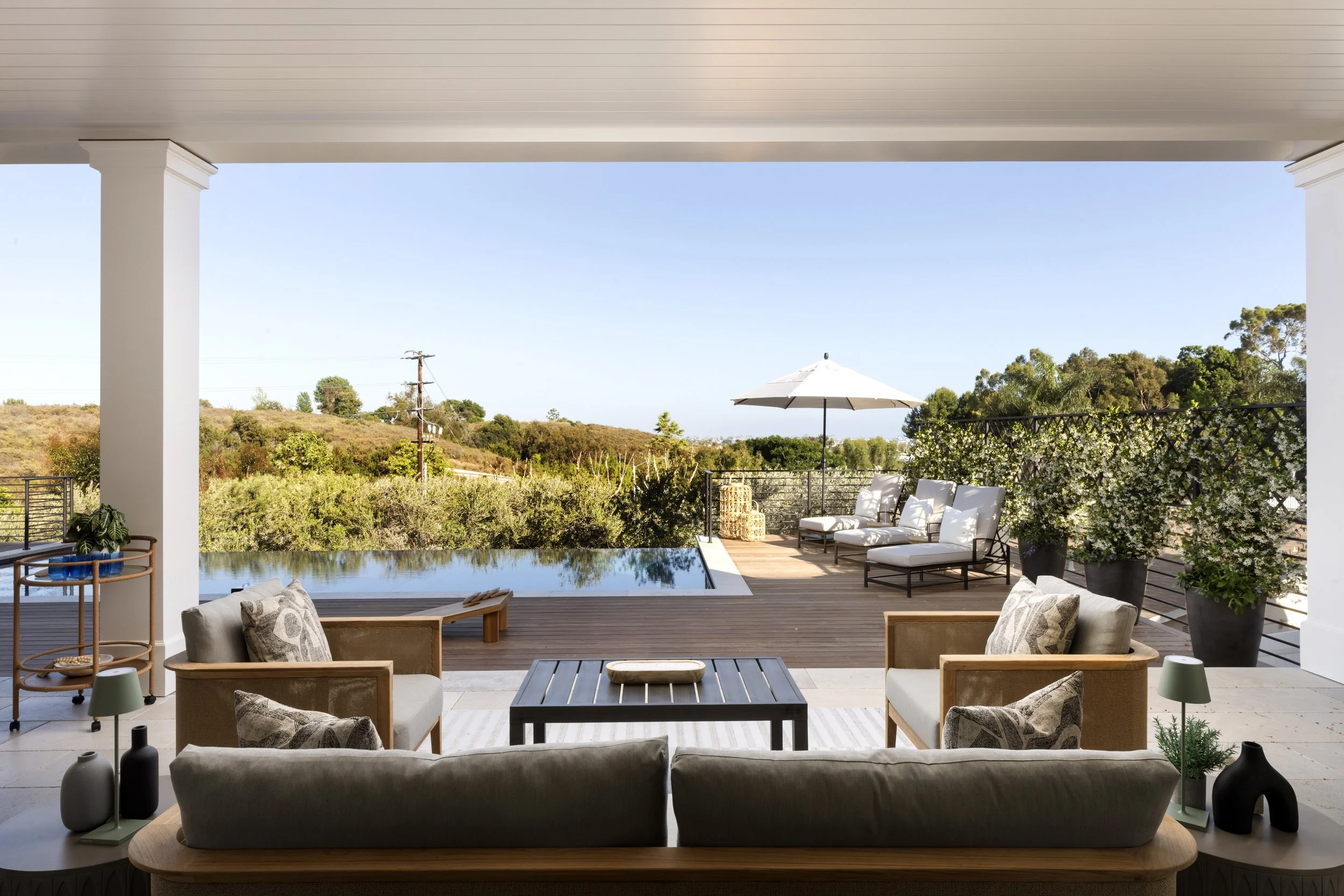 Outdoor patio with seating area including sofas and chairs, a swimming pool, and a scenic view of trees and hills in the background under a clear blue sky.