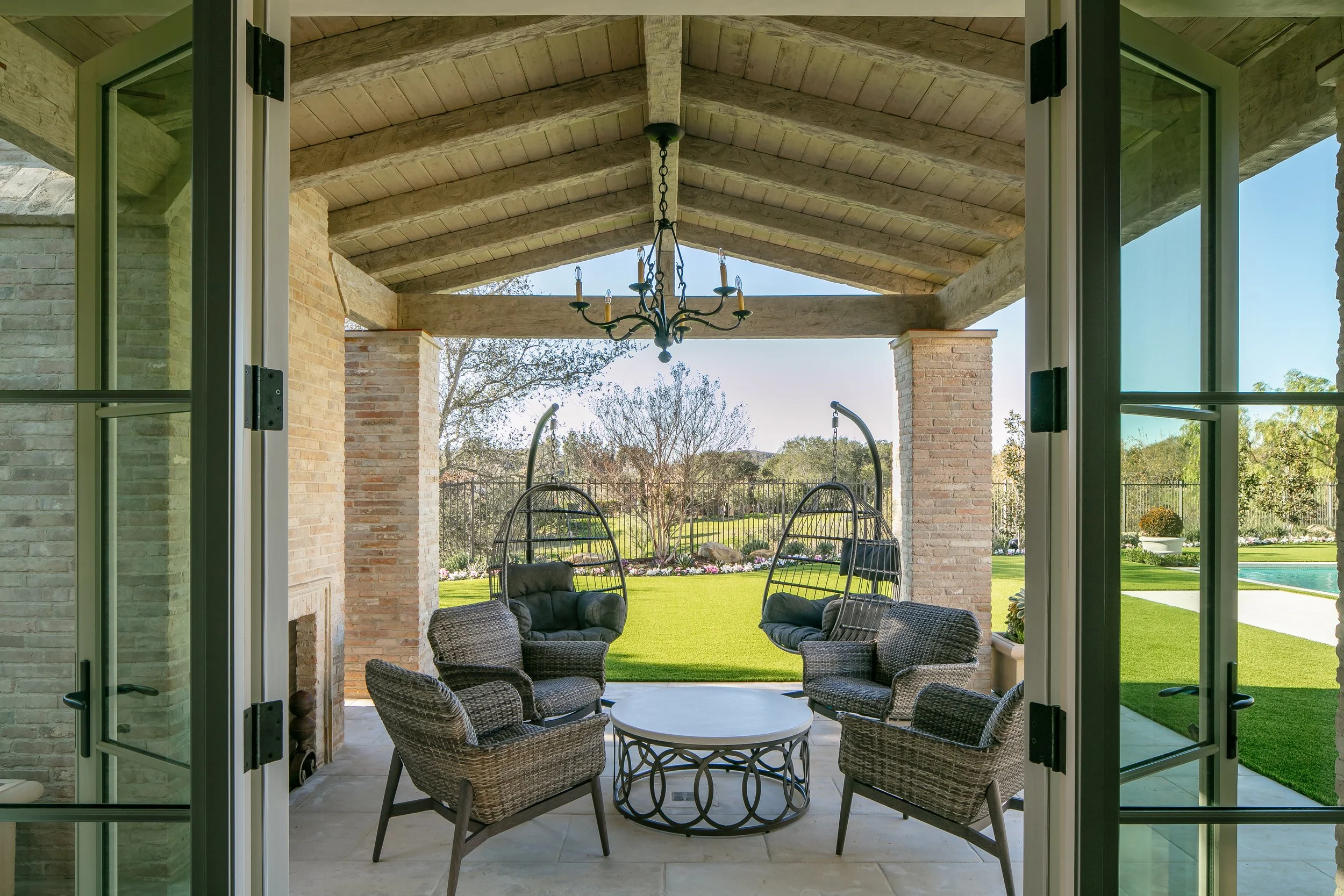 View from a screened porch looking into a backyard with outdoor seating, a chandelier hanging from a wooden beamed ceiling, trees, and a lush lawn.