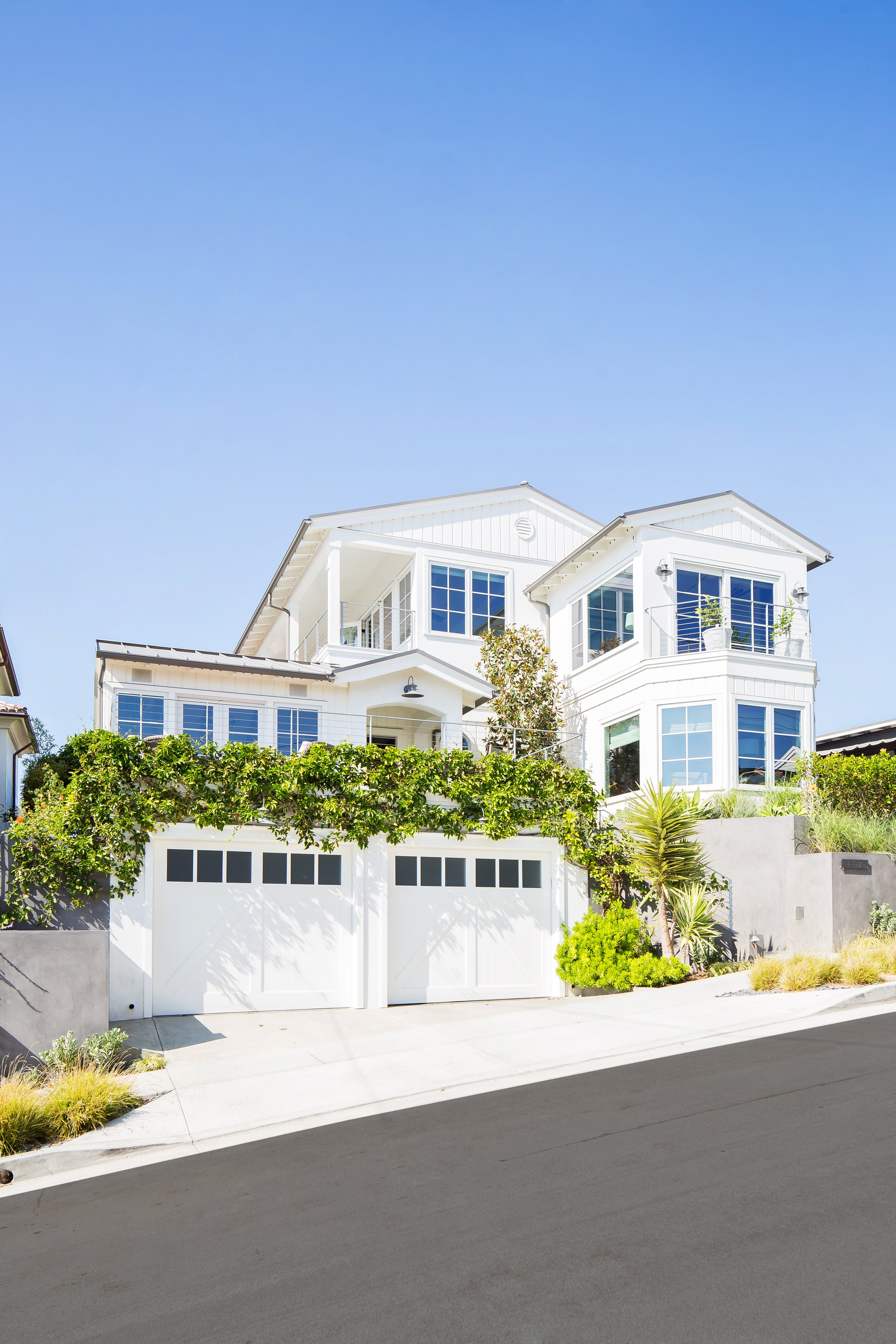 A modern white multi-story house with large windows, a garage with a white door featuring small square windows, and landscaped greenery including trees, shrubs, and climbing plants, on a sunny day with a clear blue sky.