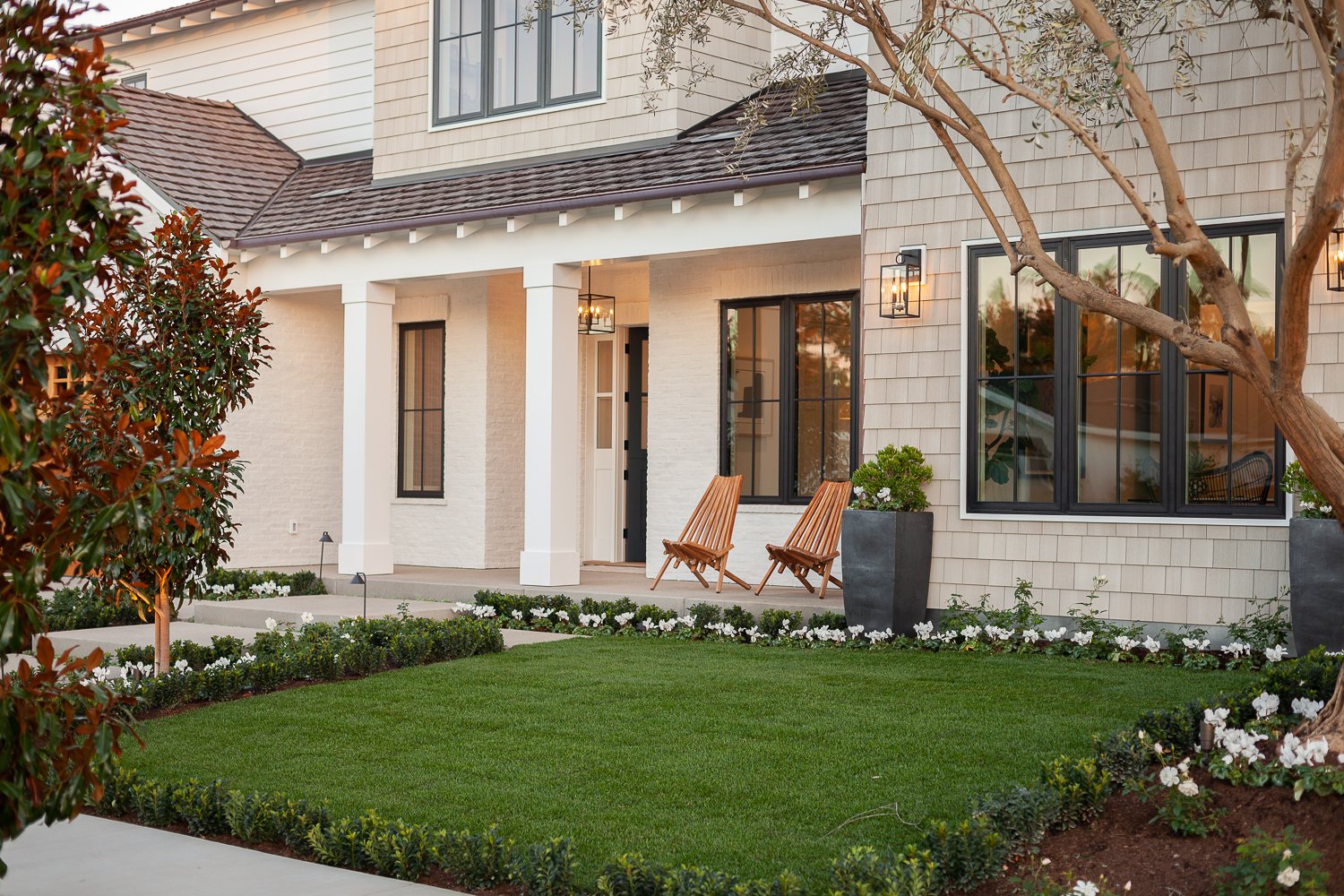 Front porch of a modern house with a well-manicured lawn, two wooden chairs, potted plants, and large windows reflecting the sky.