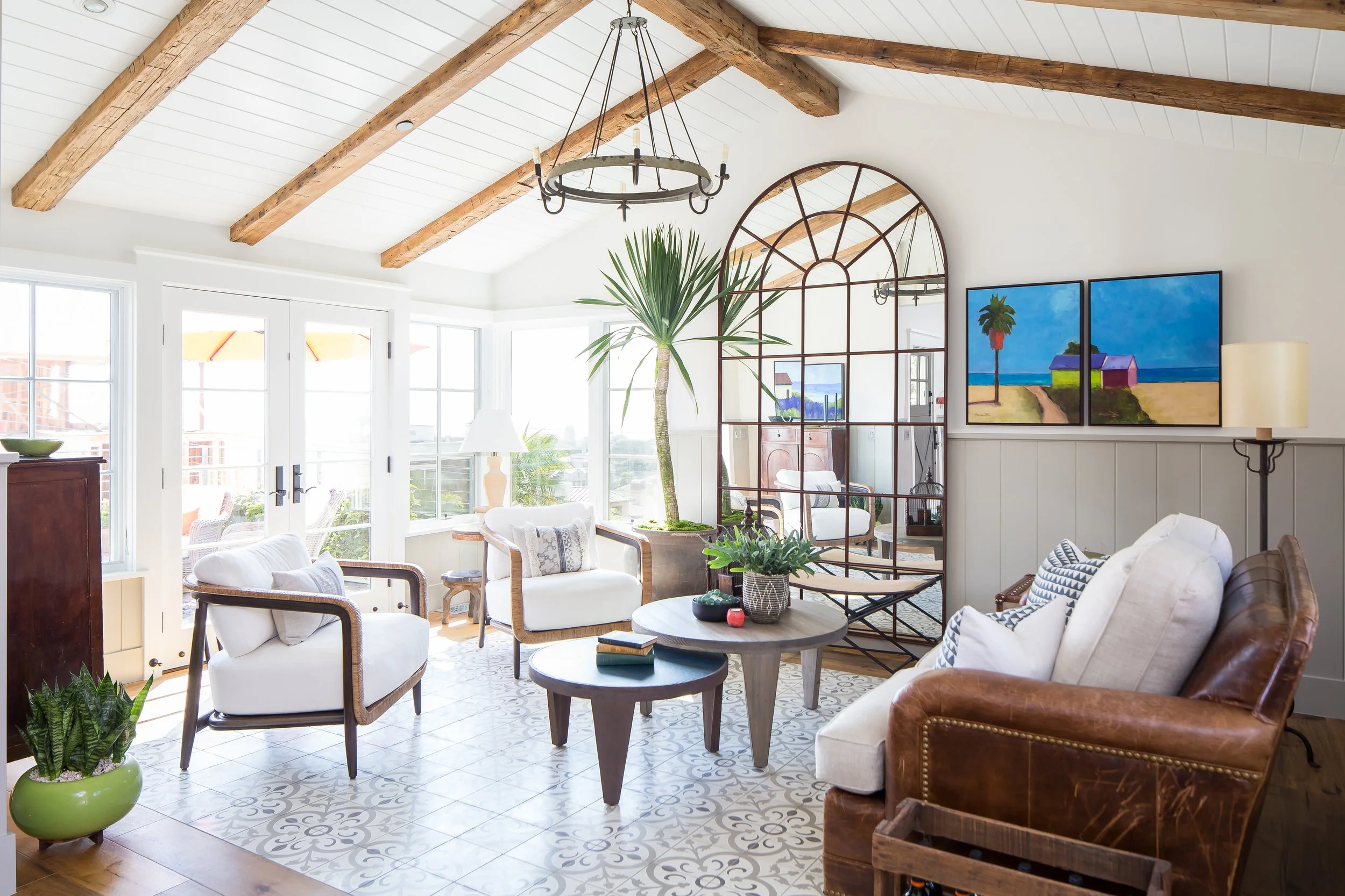A bright living room with large glass doors and windows letting in natural light. The room features a vaulted ceiling with exposed wooden beams, a circular wrought iron chandelier, and a large mirror with an arched metal frame. There are multiple sea