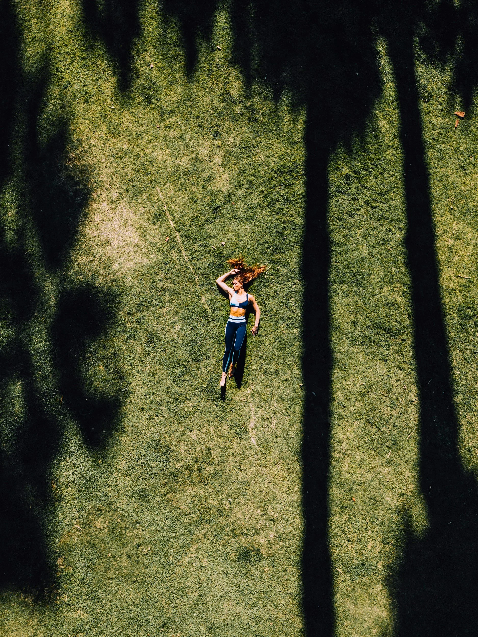 Athena Grace Foundation Experience, A woman lying on grass with her head tilted back, eyes closed, and hair spread out, surrounded by shadows cast by trees.