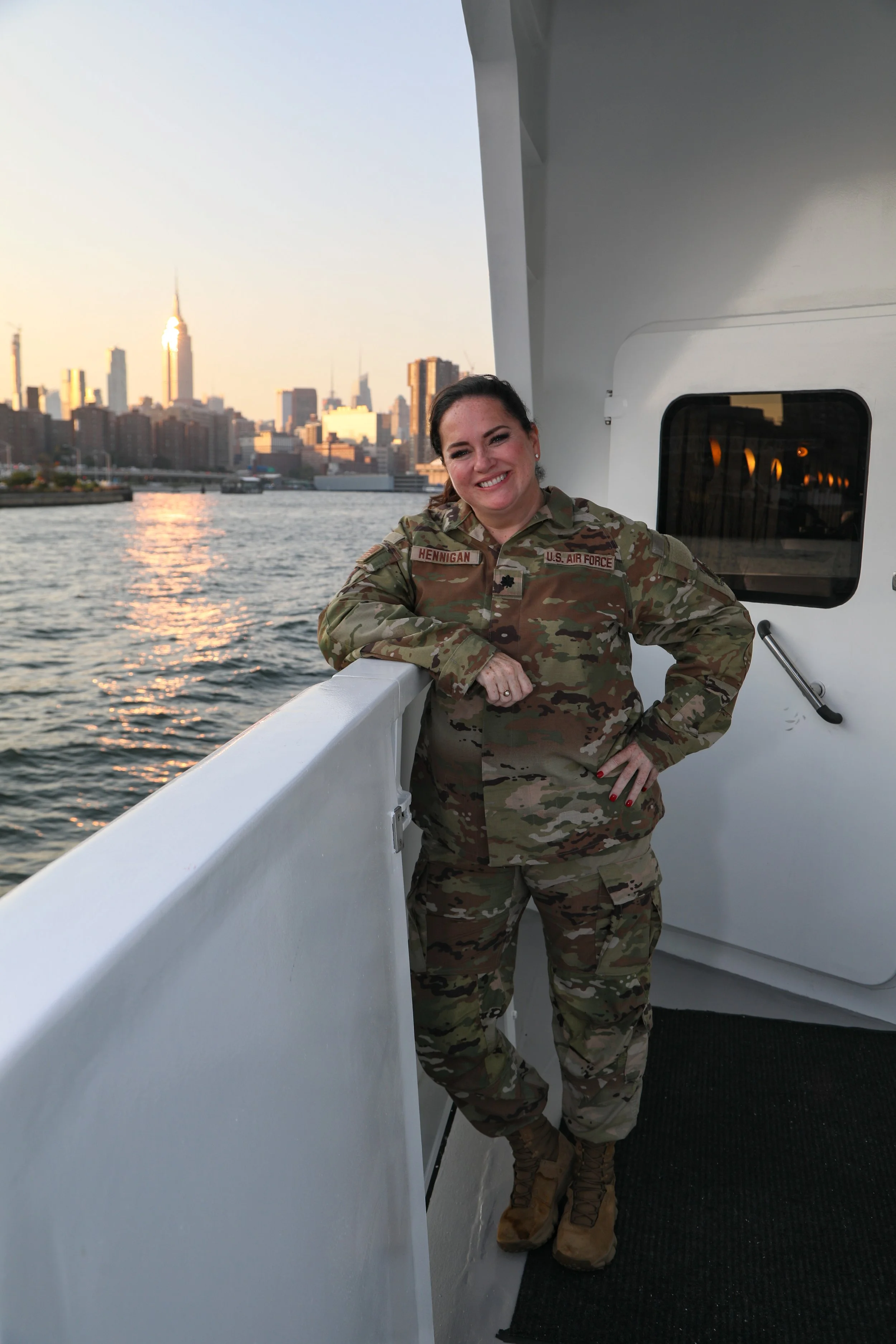 Karen Herrigan, founder of Athena Grace Foundation  in military camouflage uniform standing on a boat with a city skyline in the background, smiling at the camera during sunset.
