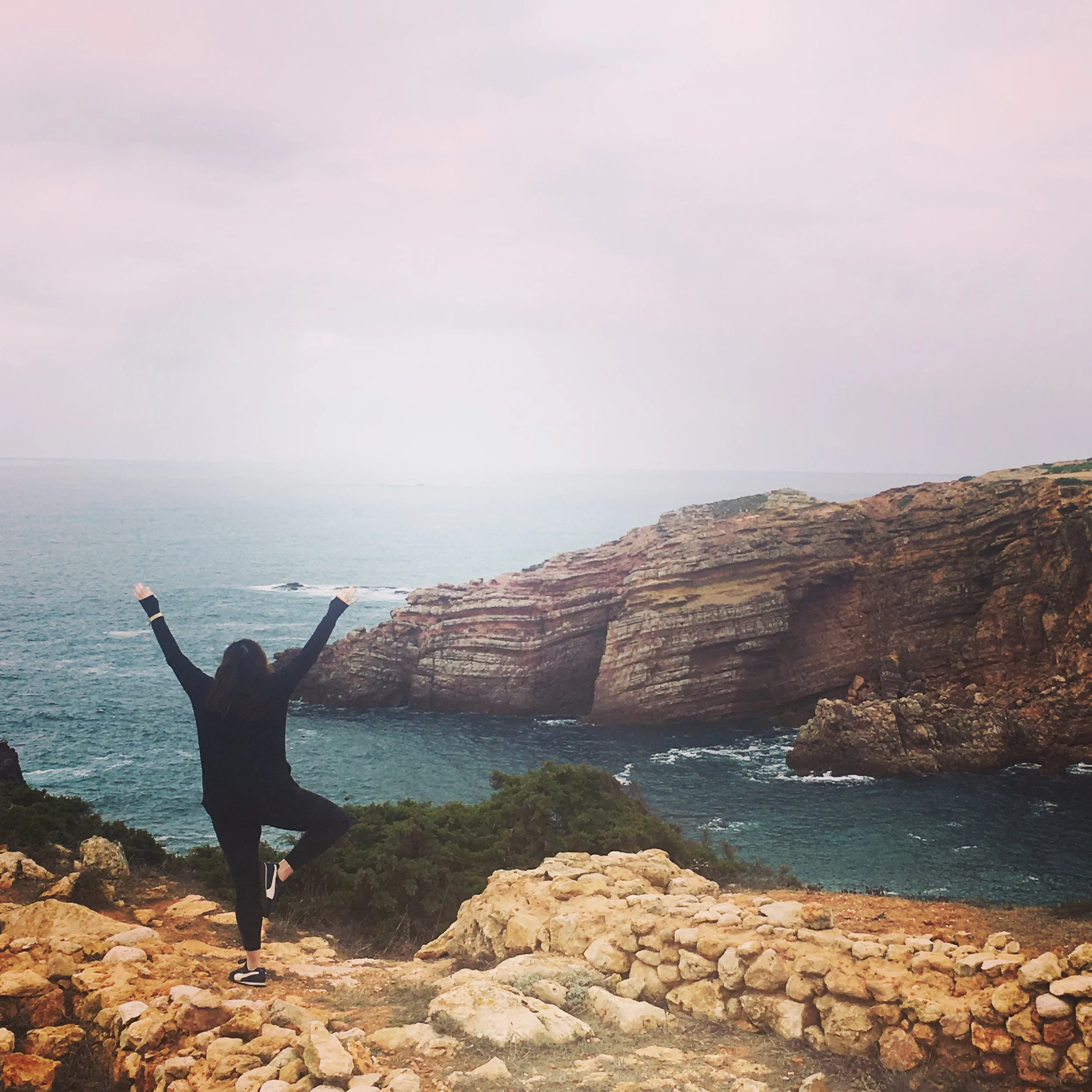 Athena Grace Foundation, a person standing on rocky terrain near the coast with arms raised, overlooking the ocean and cliffs under a cloudy sky.