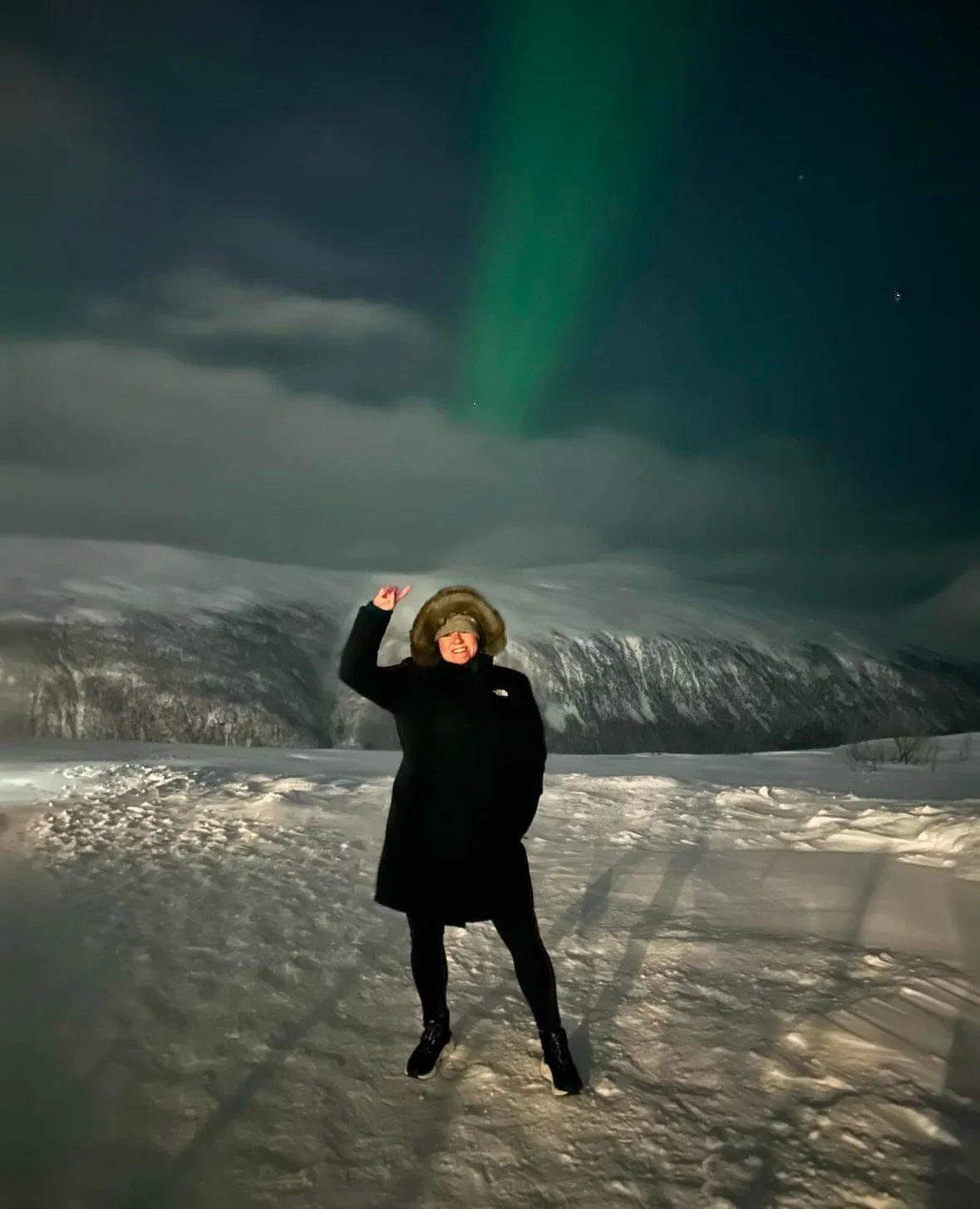 Karen Herrigan, founder of Athena Grace Foundation wearing winter clothing standing on snowy landscape with northern lights visible in the night sky.