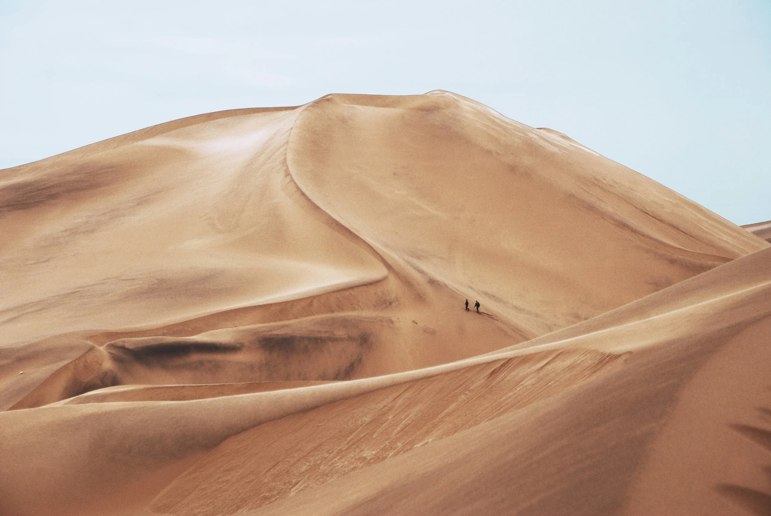 Athena Grace Lifetime Adventure, nominate a Goddess. Two people walking in a vast desert with large sand dunes and a pale blue sky.
