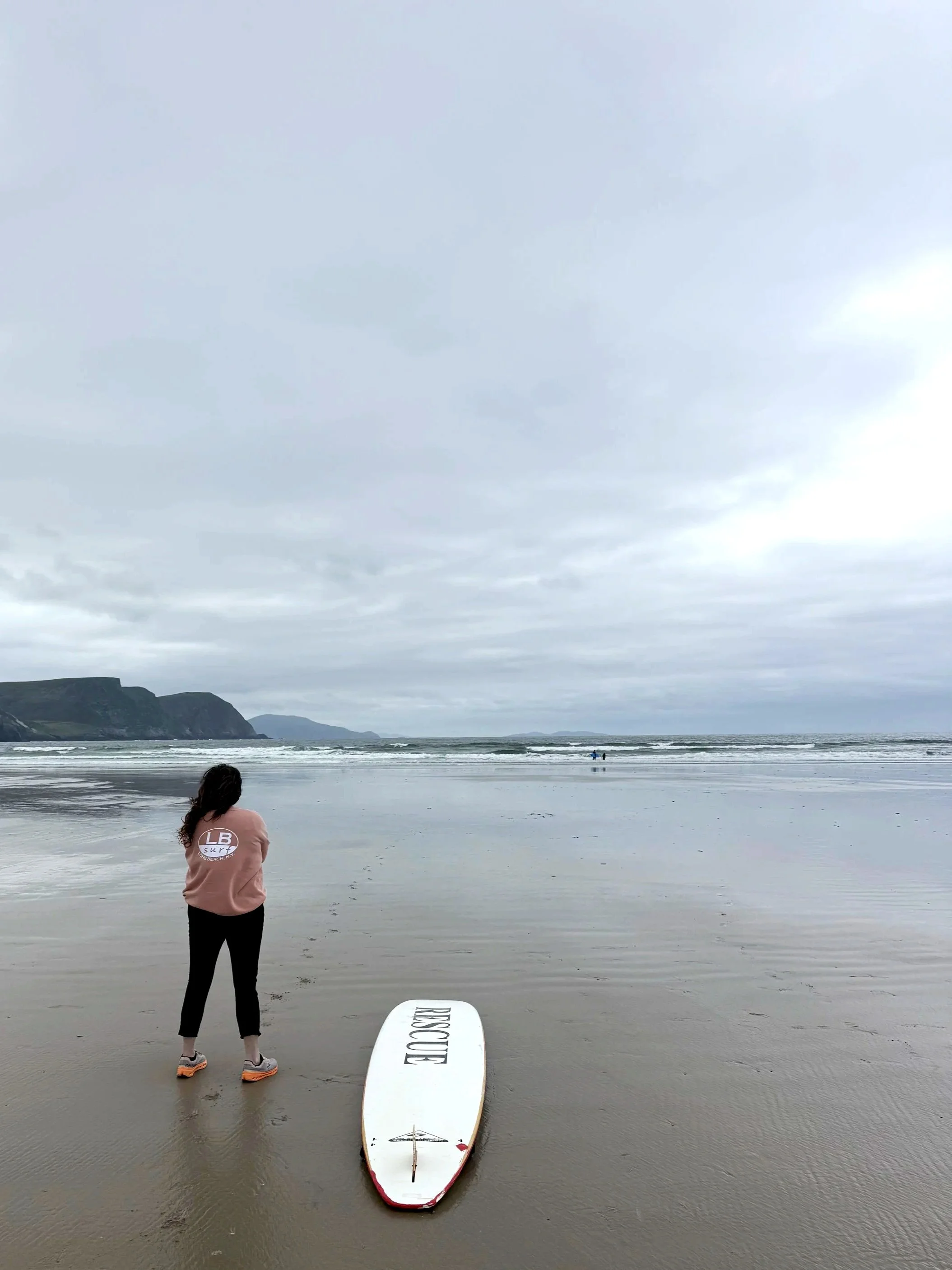 Karen Herrigan, founder of Athena Grace Foundation standing on a beach facing the ocean, with a surfboard on the sand in front of them. The sky is cloudy and there are cliffs in the distance.