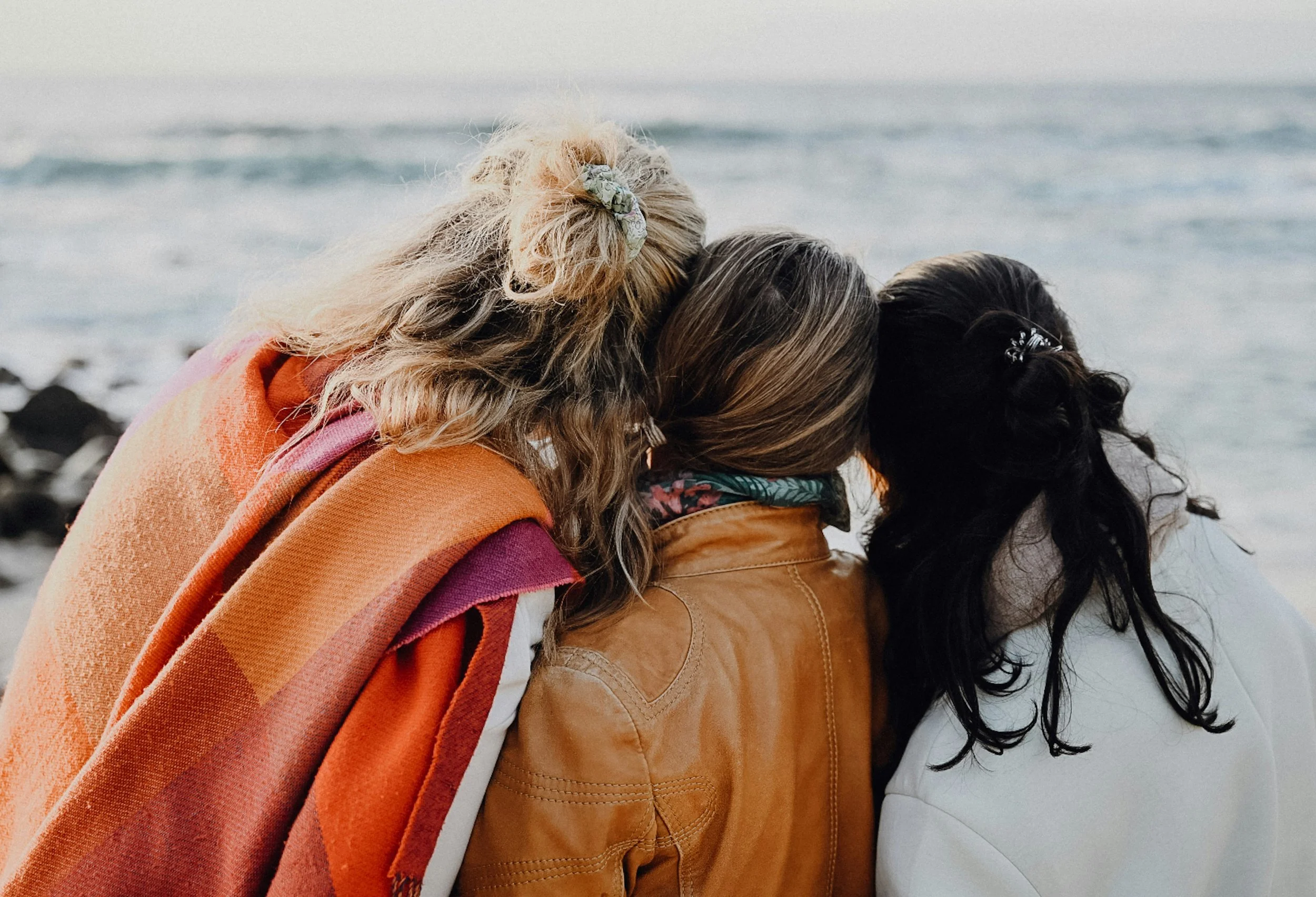 Athena Grace Gatherings: Three women with different hair colors and styles, wearing warm clothing, are sitting by the water and looking out at the ocean.