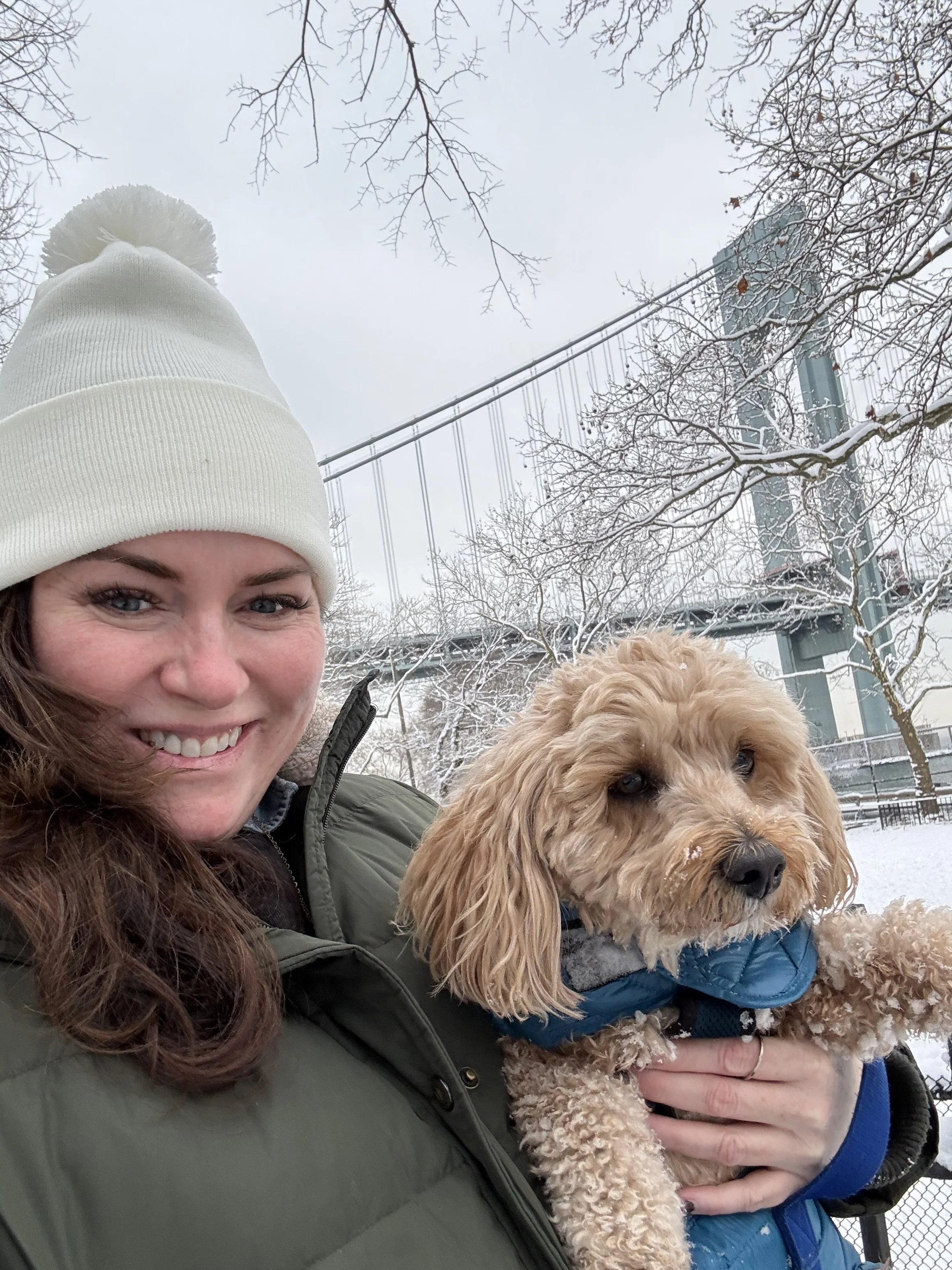 Karen Herrigan, founder of Athena Grace Foundation smiling in a white winter hat holding a curly-haired dog in a blue coat outdoors on a snowy day with trees and a bridge in the background.