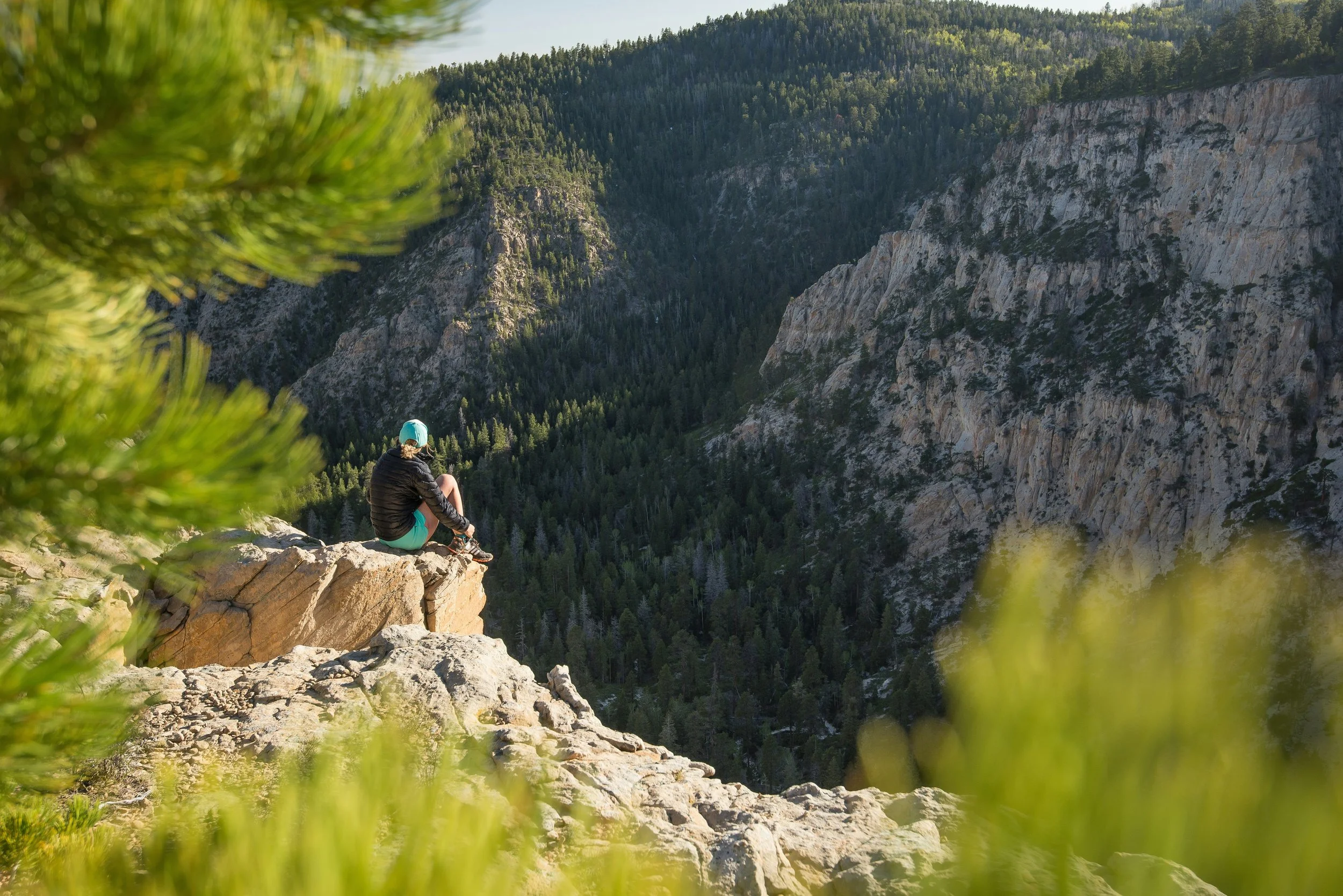 Athena Grace Nominate a Goddess for an opportunity to give them something special they serve for their service . Woman sitting on a rocky ledge overlooking a valley with dense forest and steep cliffs, framed by green foliage in the foreground.