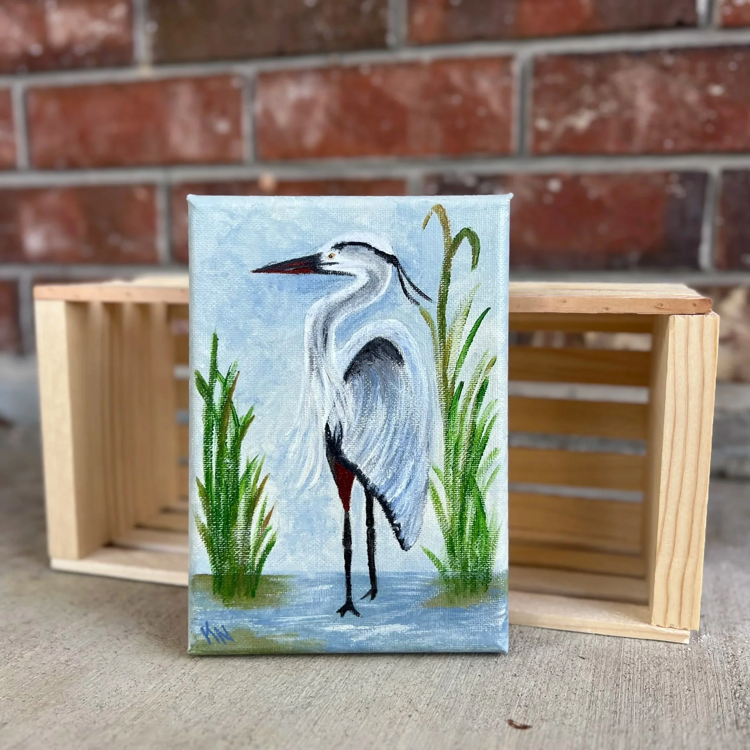 Painting of a heron standing among green reeds, on a small canvas placed on a wooden table with a brick wall in the background.