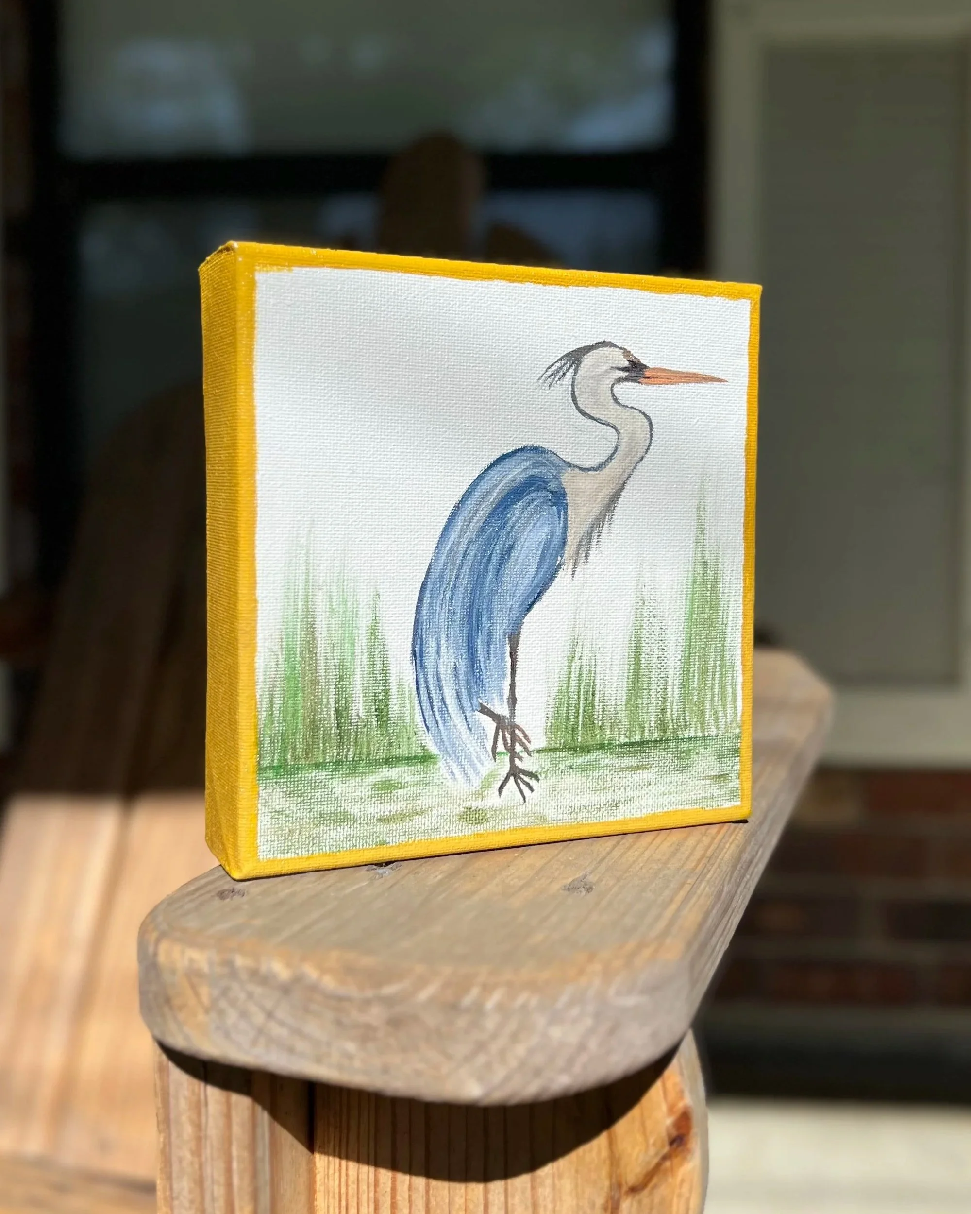 A small rectangular canvas with a yellow border features a watercolor painting of a heron standing among green grass.