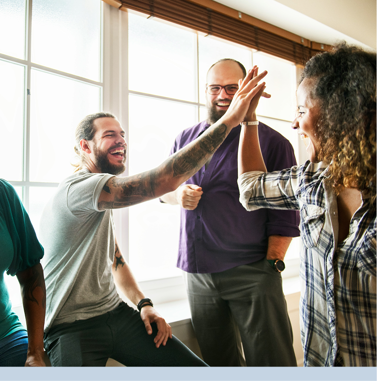 Group of four diverse people high-fiving and smiling in a sunlit room with large windows.