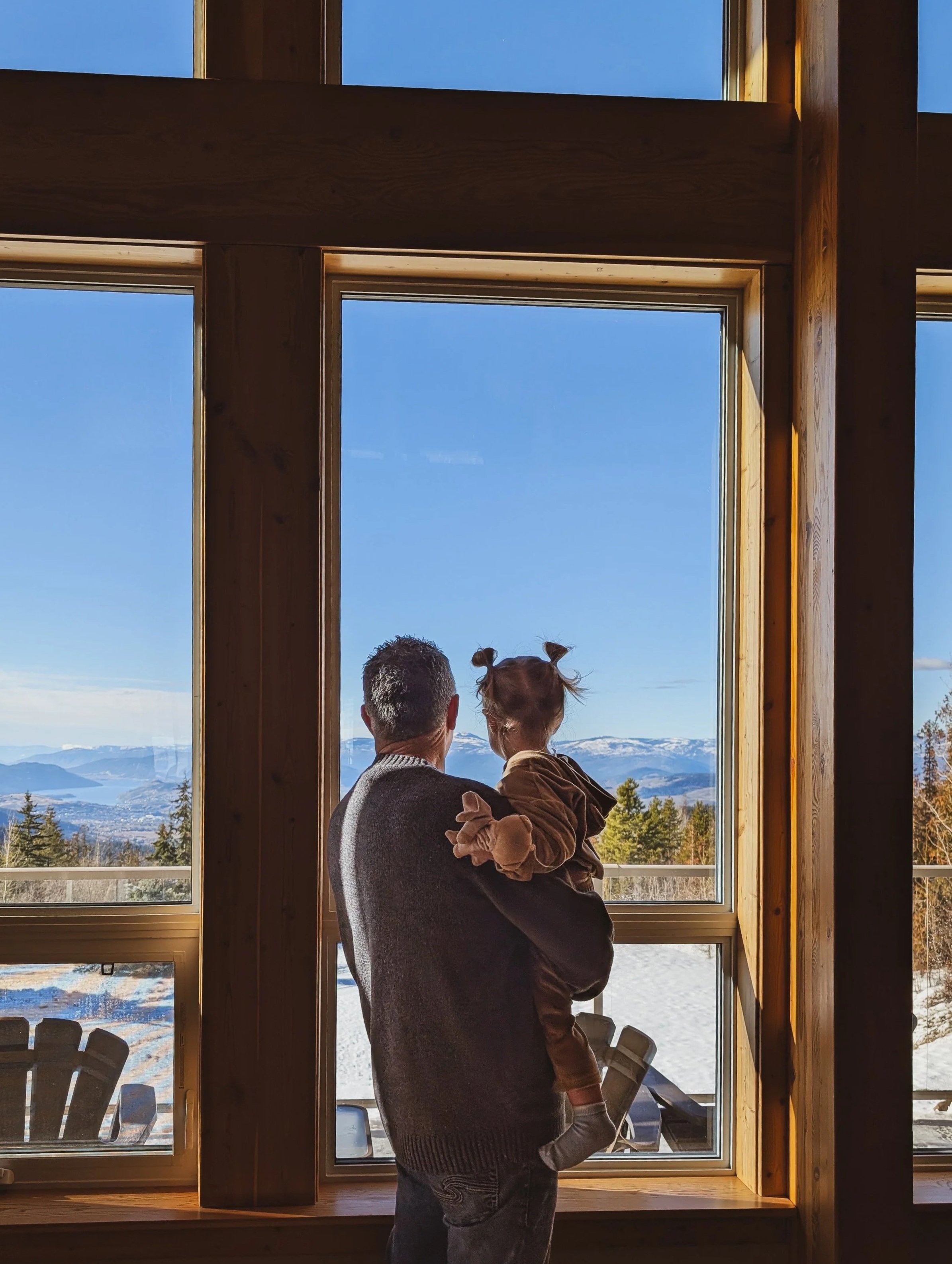 A man with short, grey hair holding a young girl with two pigtails and light brown hair, looking out a large window showing a hilly landscape with snow and forests during daytime.
