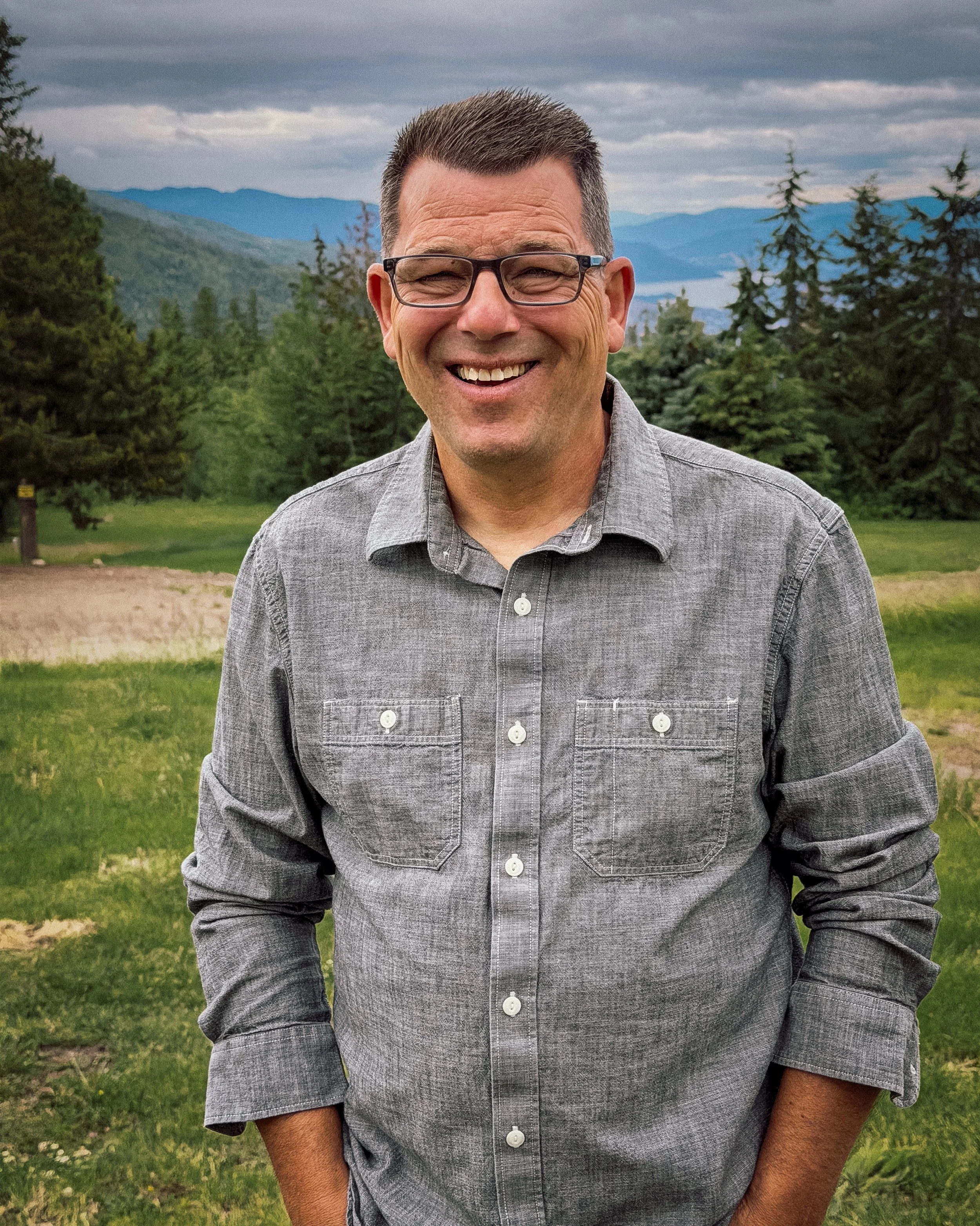 A smiling man with glasses, wearing a gray button-up shirt, standing outdoors in a lush, green landscape with trees and mountains in the background under a cloudy sky.