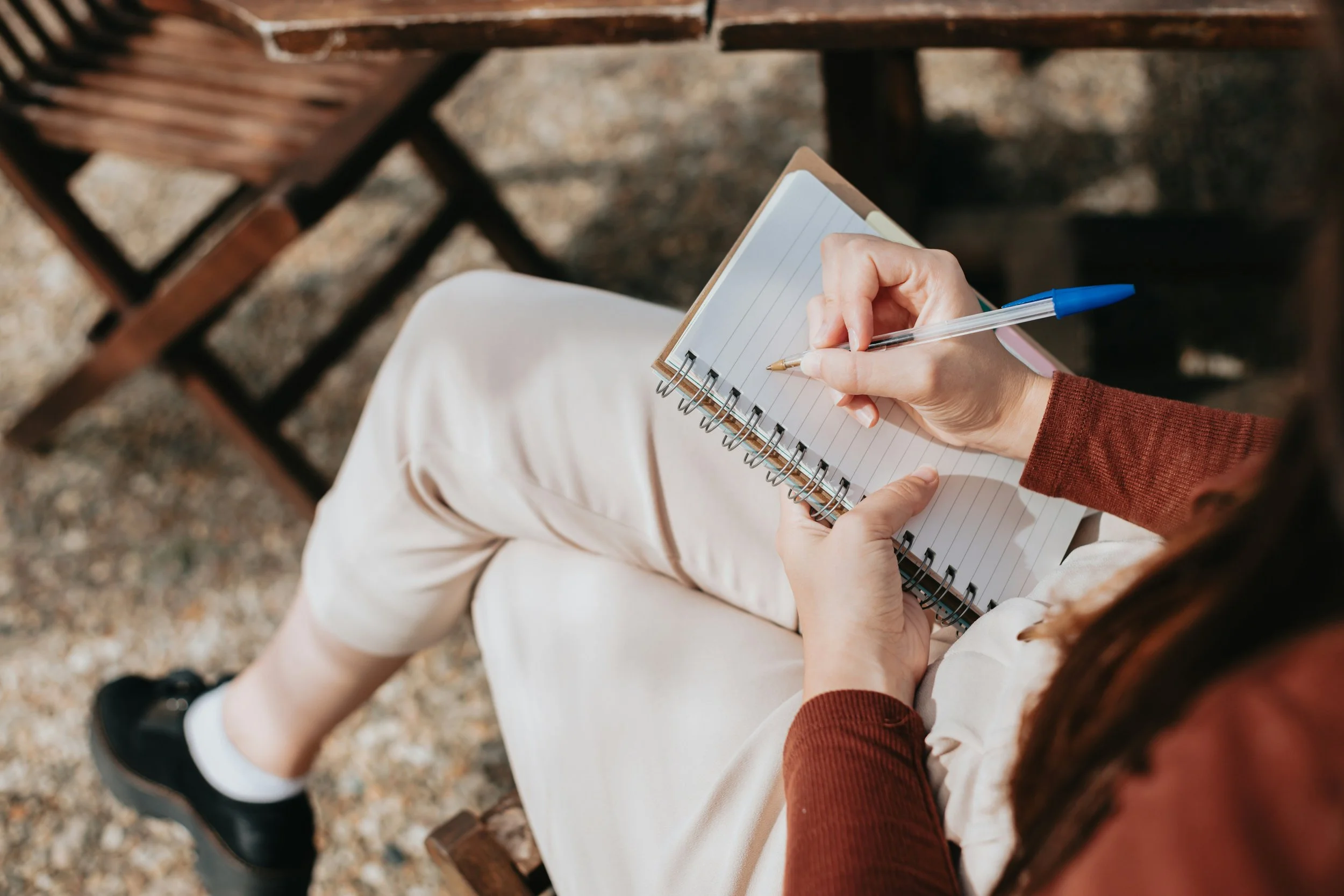 A person sitting on a wooden bench outdoors, writing in a small spiral notebook with a pen, wearing light-colored pants and a brown long-sleeve shirt.
