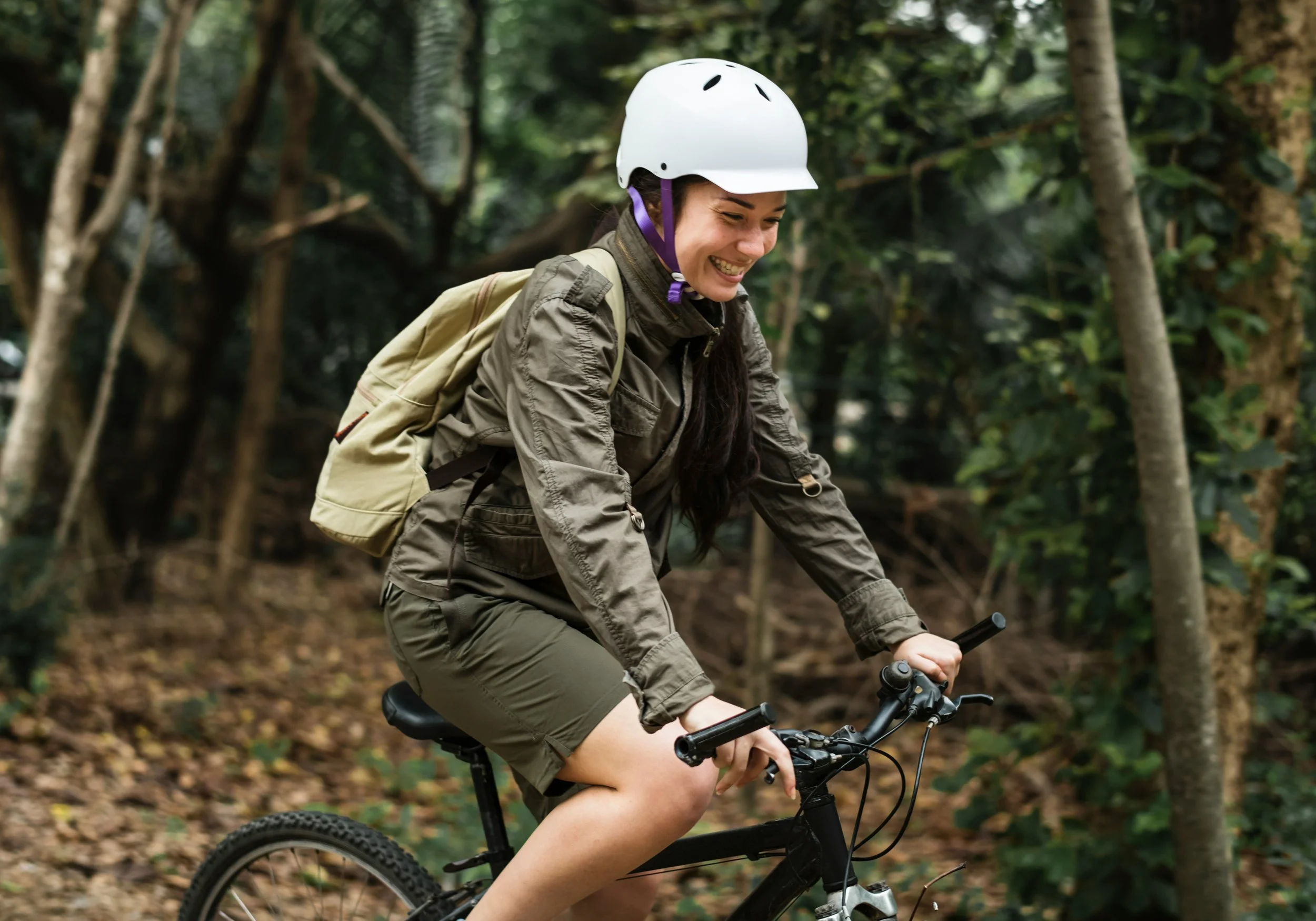 Woman wearing a white helmet riding a mountain bike through a forest trail, smiling and enjoying the outdoor adventure.