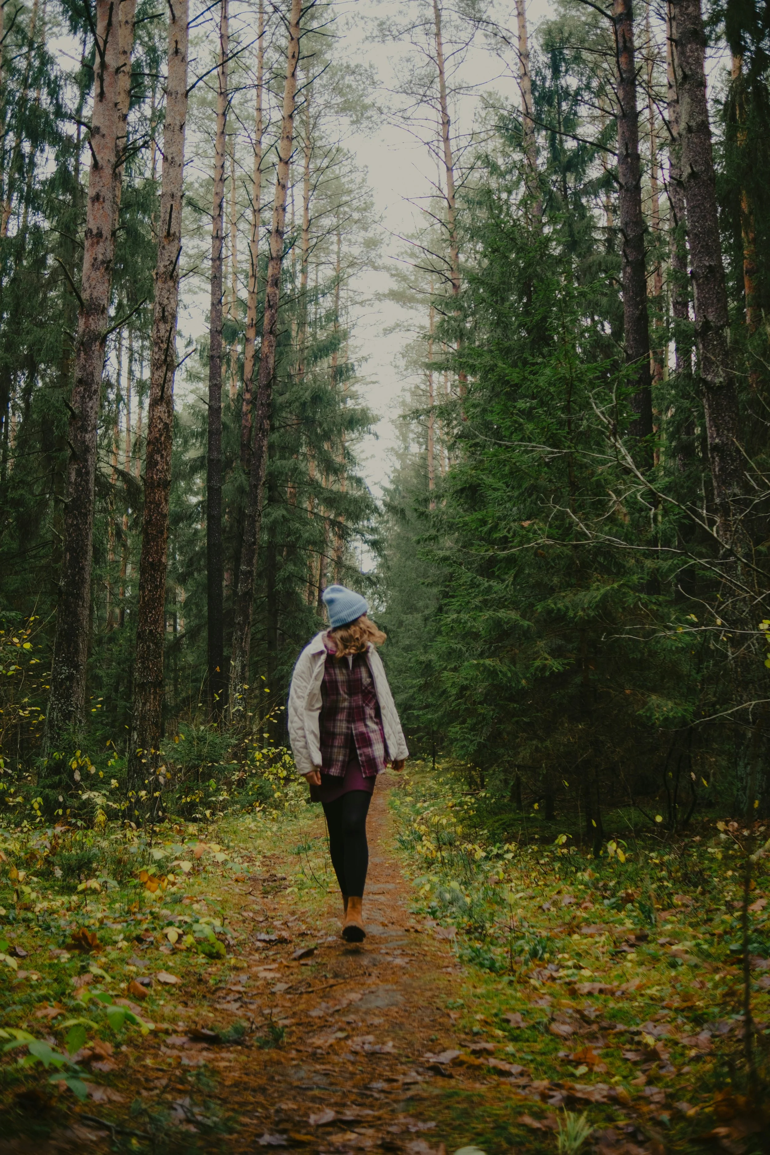 A person with curly hair walking along a forest trail in autumn, wearing a gray beanie, beige jacket, plaid shirt, and black tights.