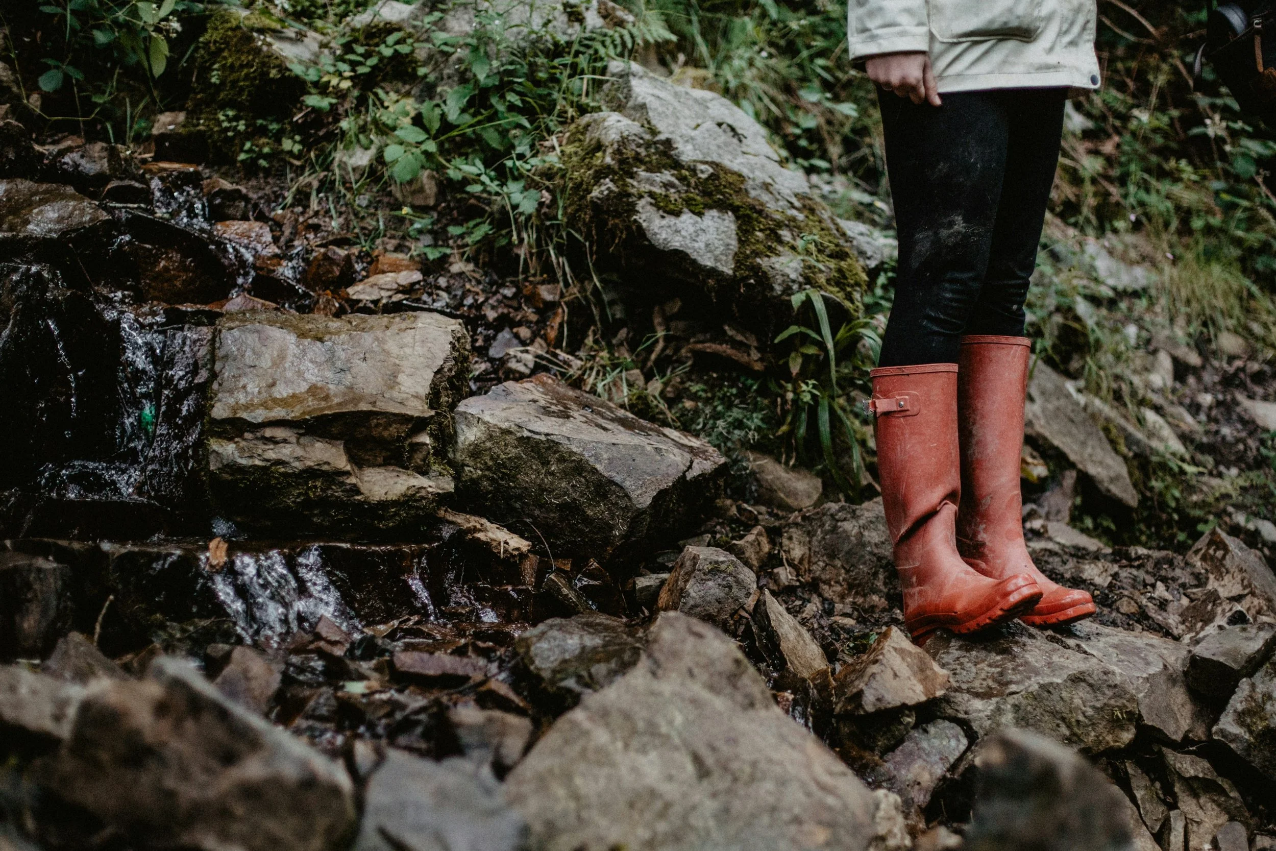 Person wearing red rubber boots standing on rocky and muddy ground near a small waterfall or stream in a forested area.