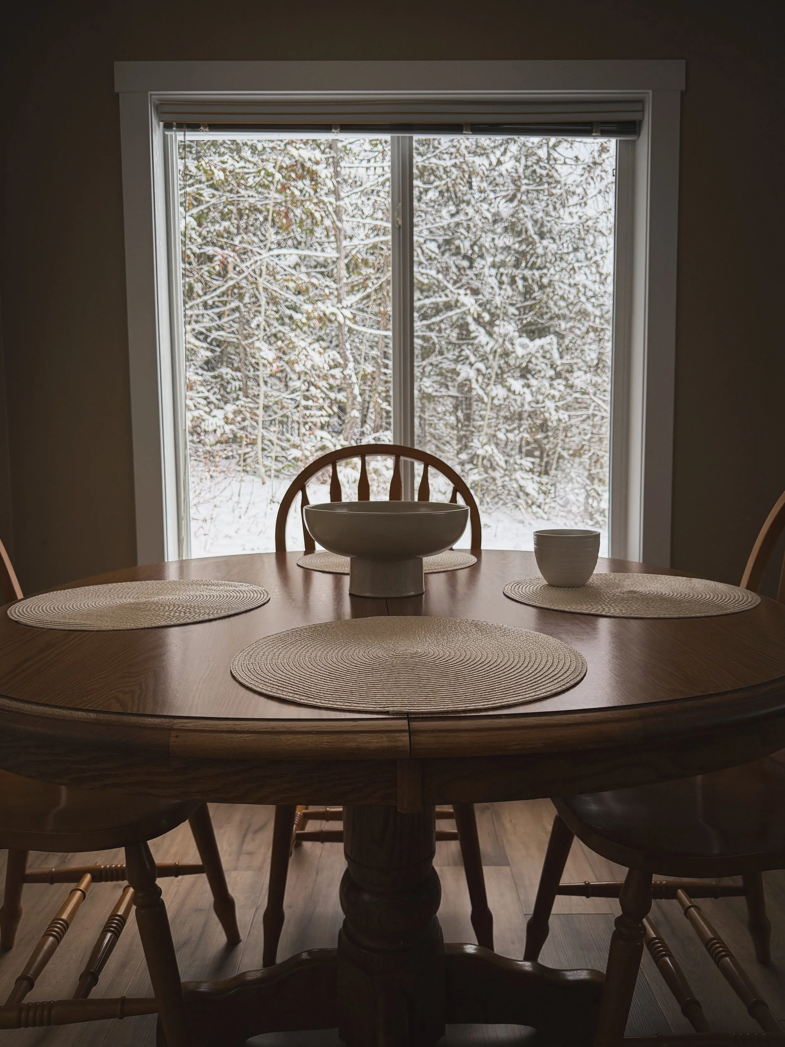 Dining room with a round oak table and a snowy view outside a window.