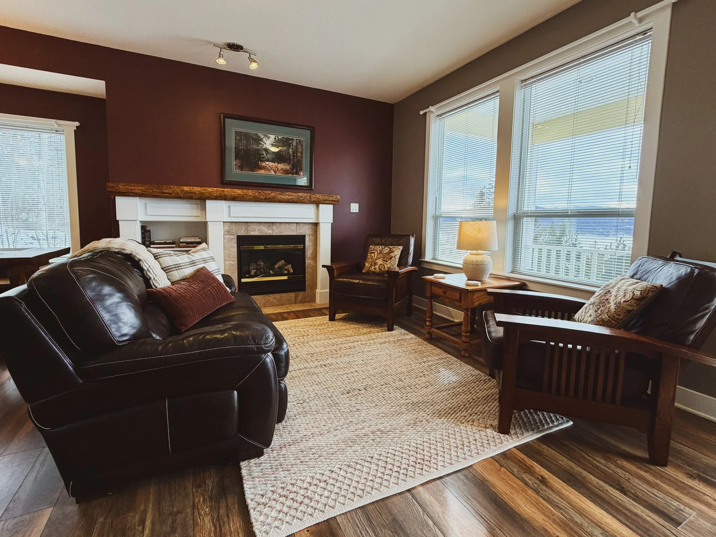 Living room with a dark leather sofa, plaid pillows, a white fireplace with a wooden mantle, a framed landscape painting, a side table with a lamp, large windows with blinds, and a rug on hardwood flooring.