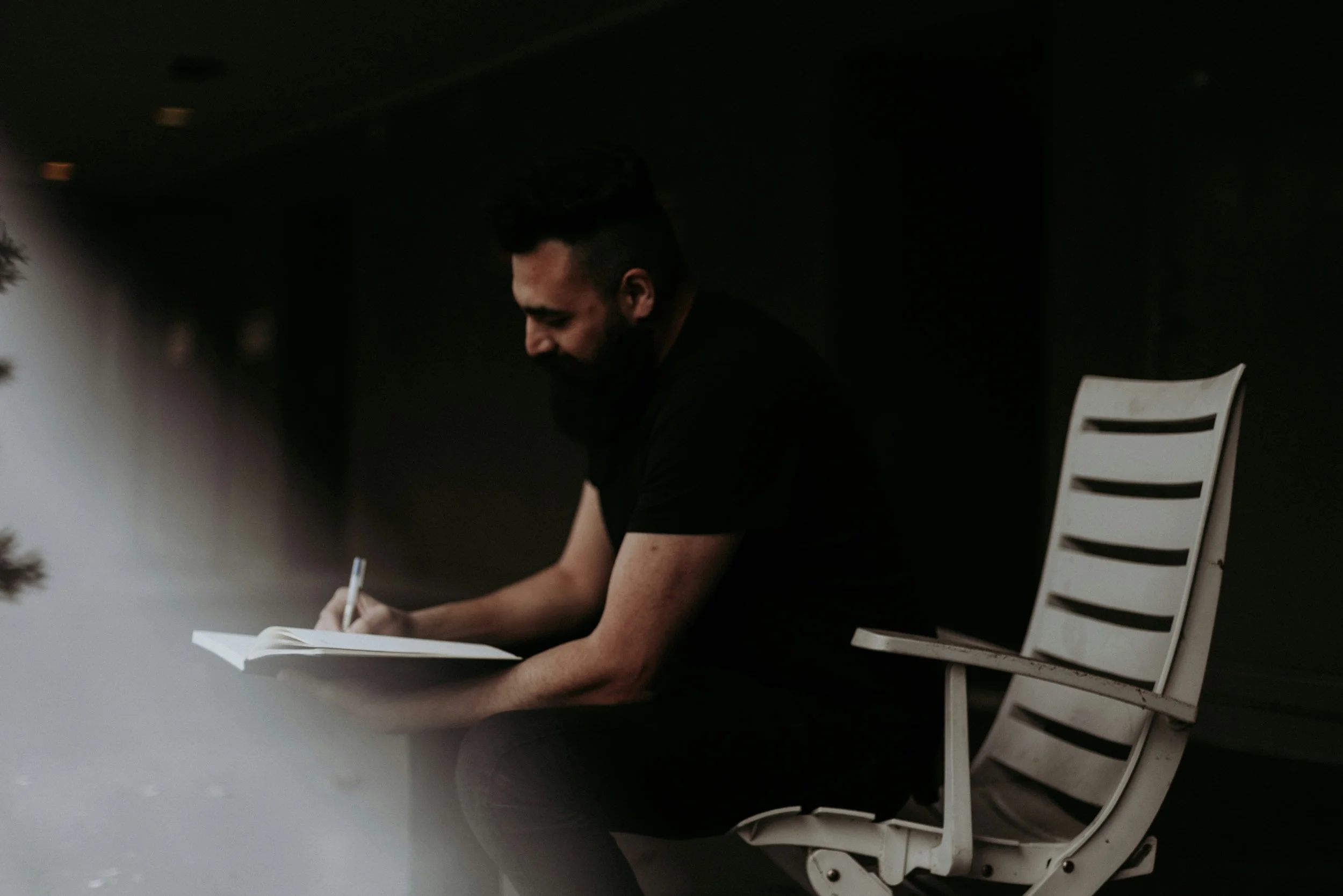A man with dark hair and a beard sitting in a white chair in a dimly lit room, writing or drawing on a piece of paper on a table.
