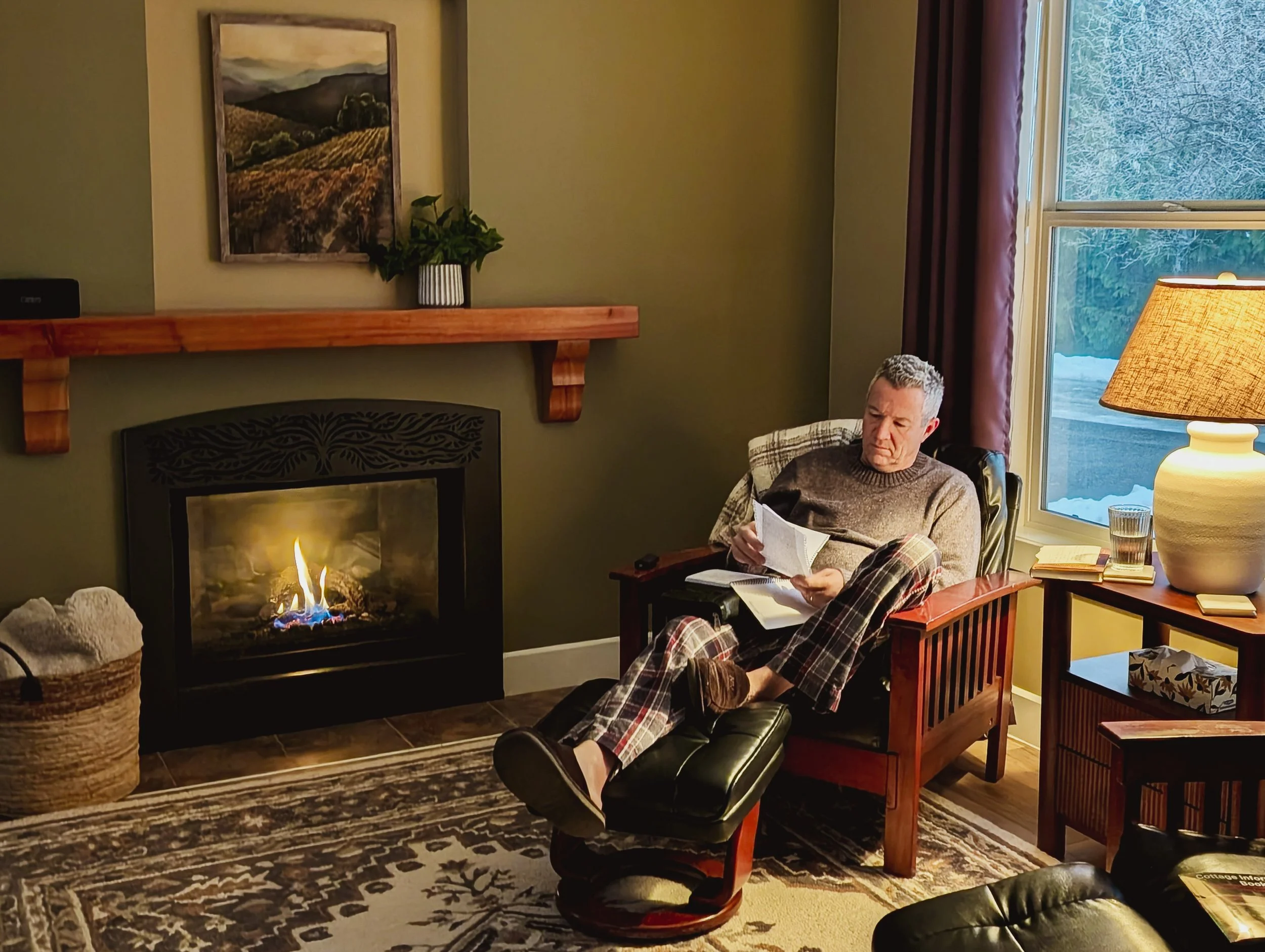 A man with gray hair and a plaid blanket sits reading a book next to a lit fireplace, a table with a glass of water, books, and a lamp, in a cozy living room with a window showing snow outside.