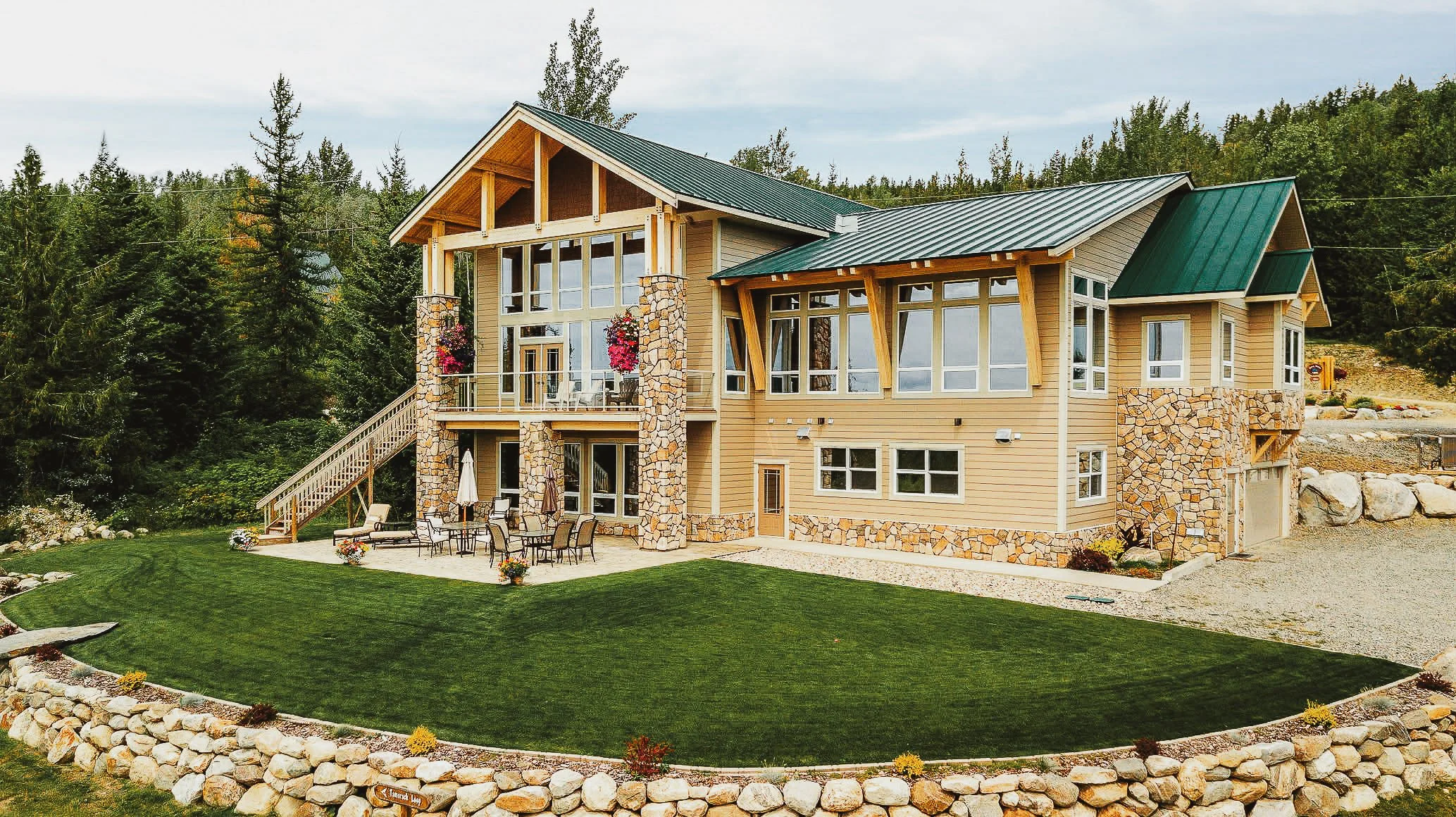 A modern two-story lodge with large windows, stone accents, green metal roof, and an outdoor patio with tables and chairs, surrounded by a landscaped yard with a stone border and greenery.
