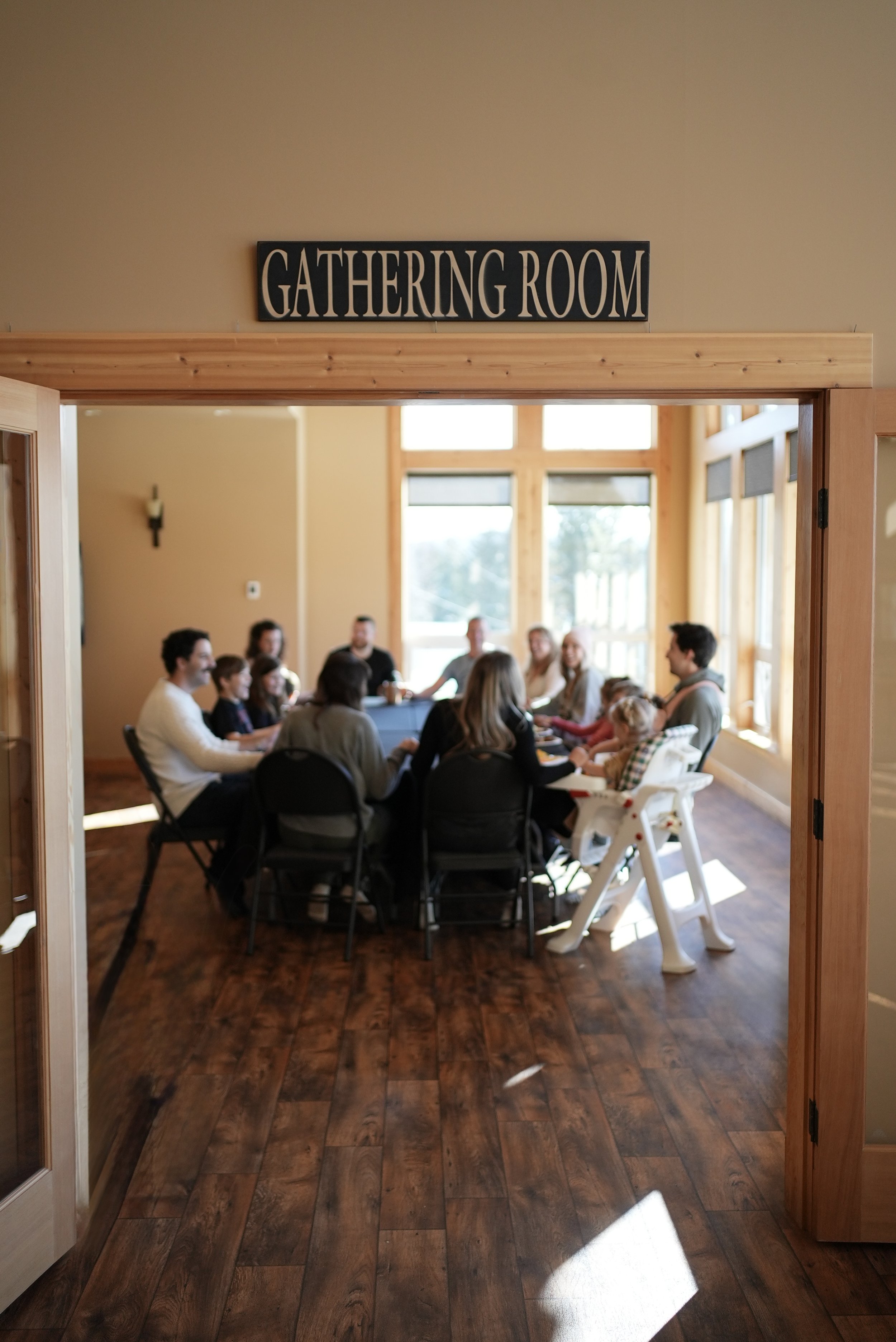 A group of people sitting around a long table in a bright, wooden room labeled 'Gathering Room' above the doorway, with natural light coming through large windows.