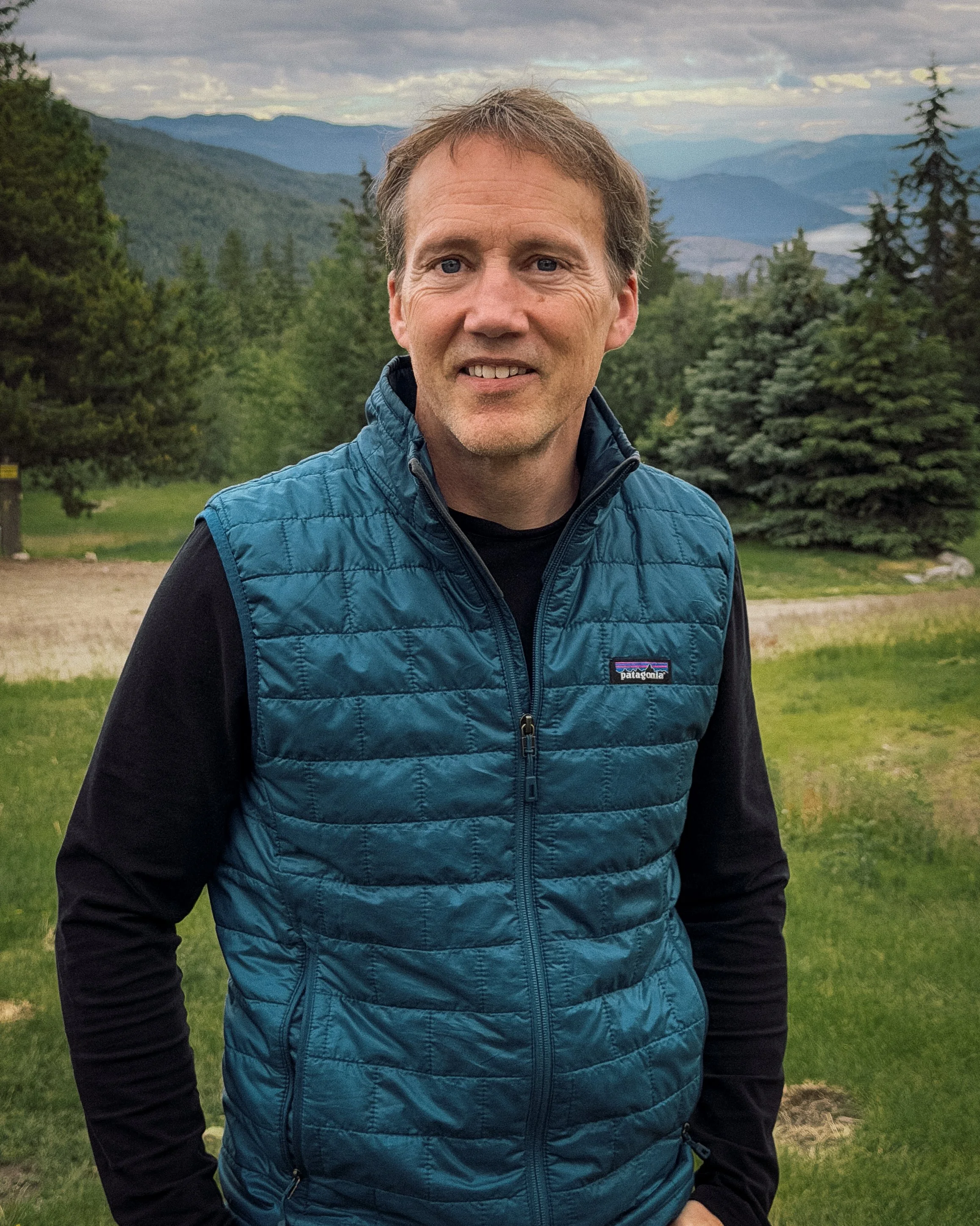 A middle-aged man with light brown hair and blue eyes, wearing a blue Patagonia vest over a black long-sleeve shirt, standing outdoors with a scenery of green trees and distant mountains under a partly cloudy sky.