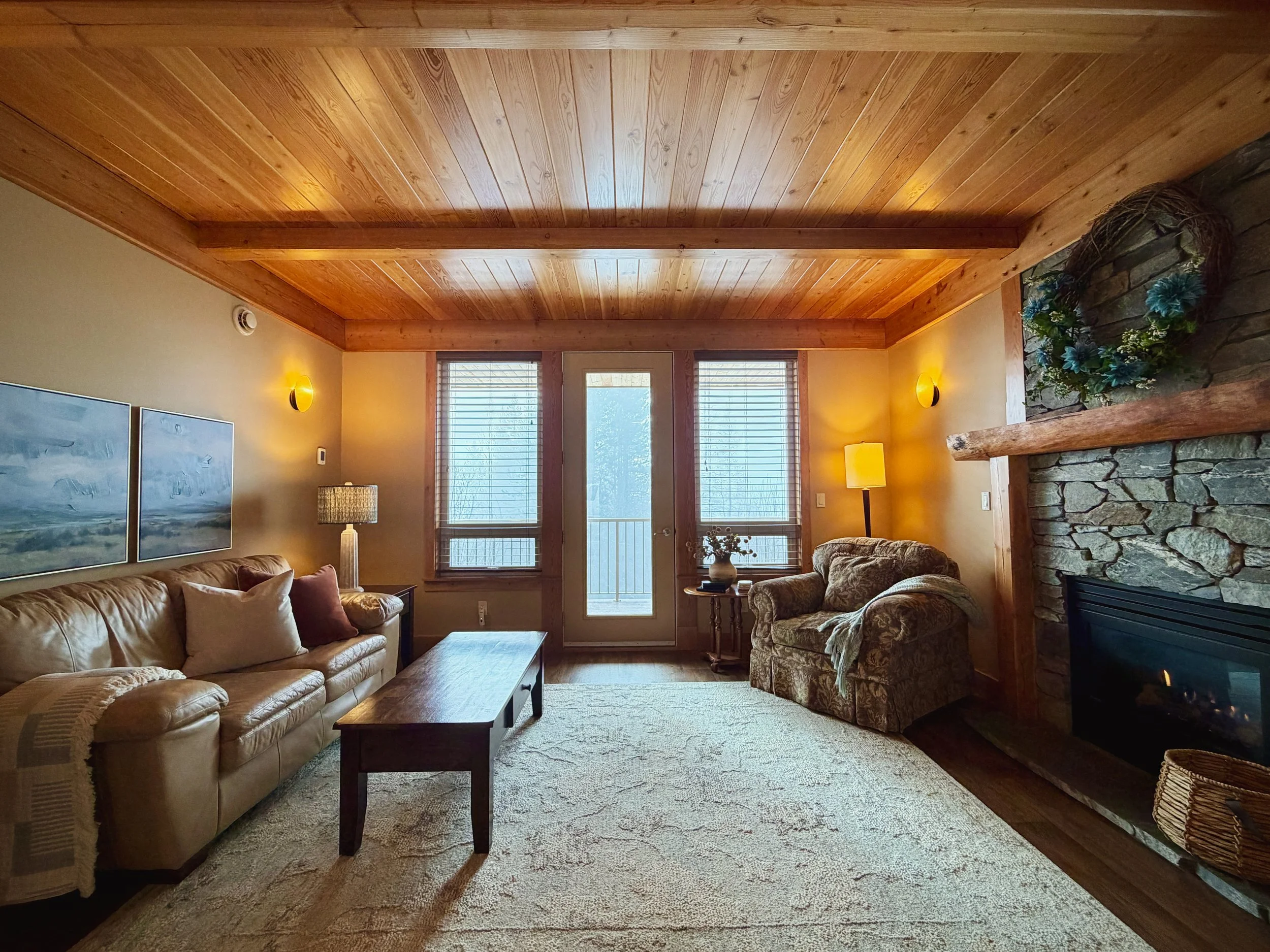 Living room with a high wood-paneled ceiling, large windows with blinds, and a stone fireplace with a wooden mantel decorated with blue and white floral arrangements. There are two sofas, a coffee table, and a lamp in the room.