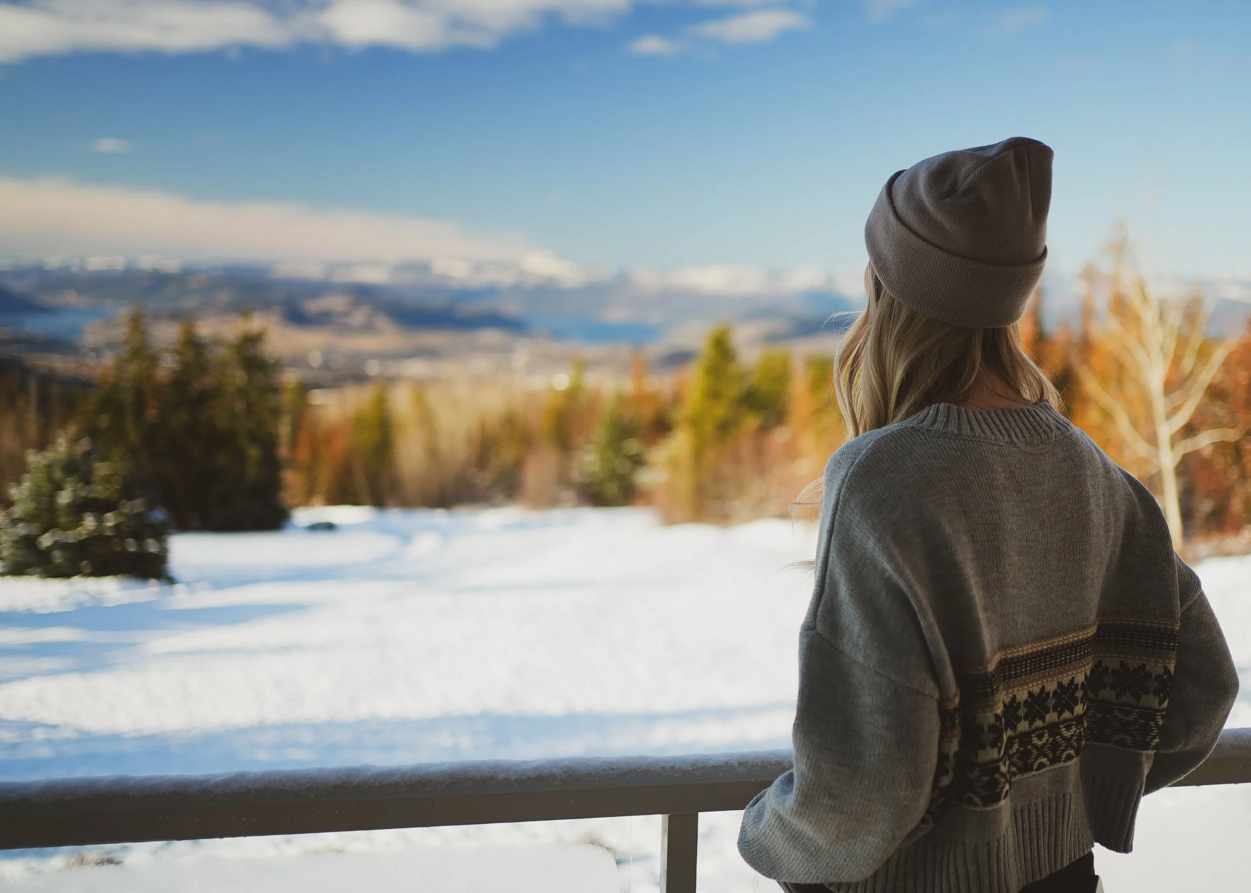 A woman with long hair wearing a gray beanie and sweater standing outdoors by a snow-covered railing, gazing at a scenic mountain landscape with snow-capped peaks and a partly cloudy sky.