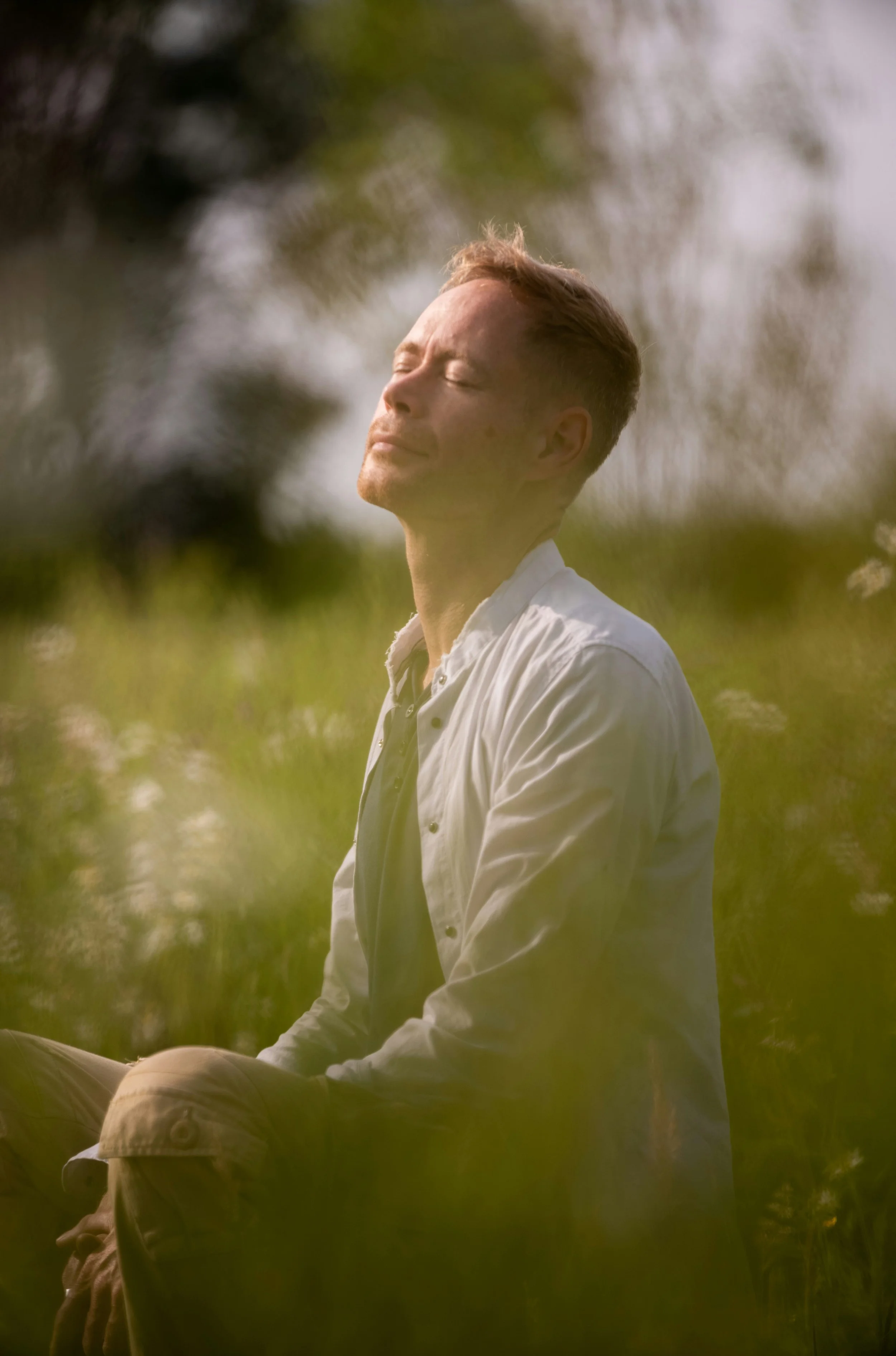 A man sitting outdoors with his eyes closed, enjoying sunlight through trees, surrounded by greenery.