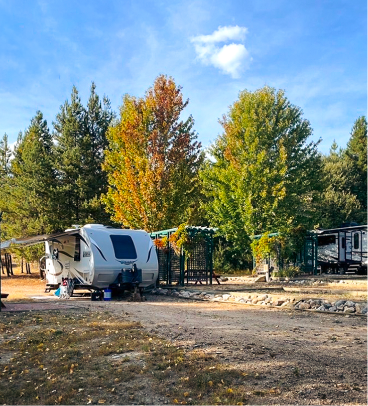 A white travel trailer parked on a dirt area with trees showing autumn colors in the background and a sunny sky with a few clouds.