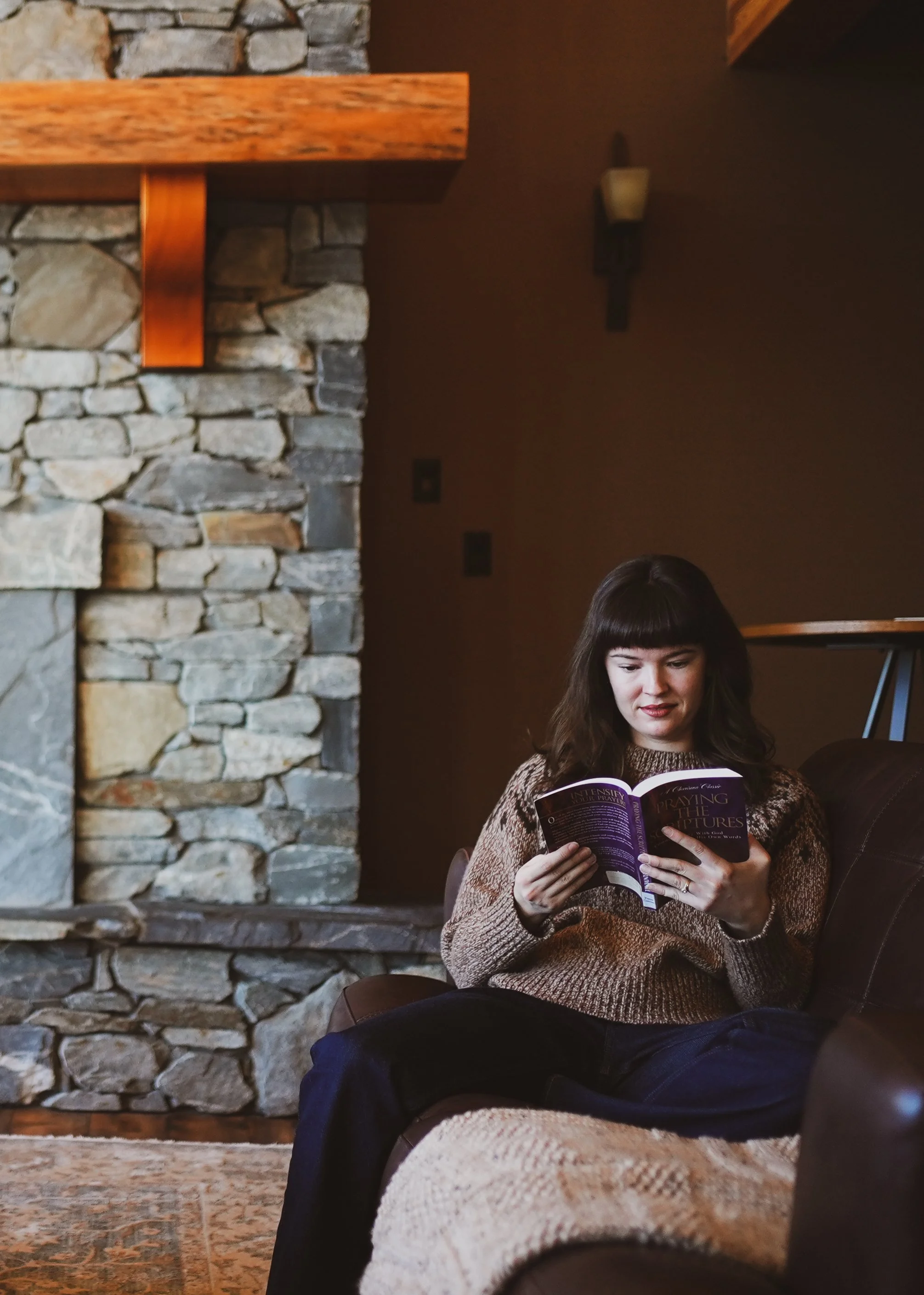 A woman with dark hair and bangs sitting on a couch, reading a magazine titled 'Praying the Pictures' in a rustic lodge with a stone fireplace and wooden beams.