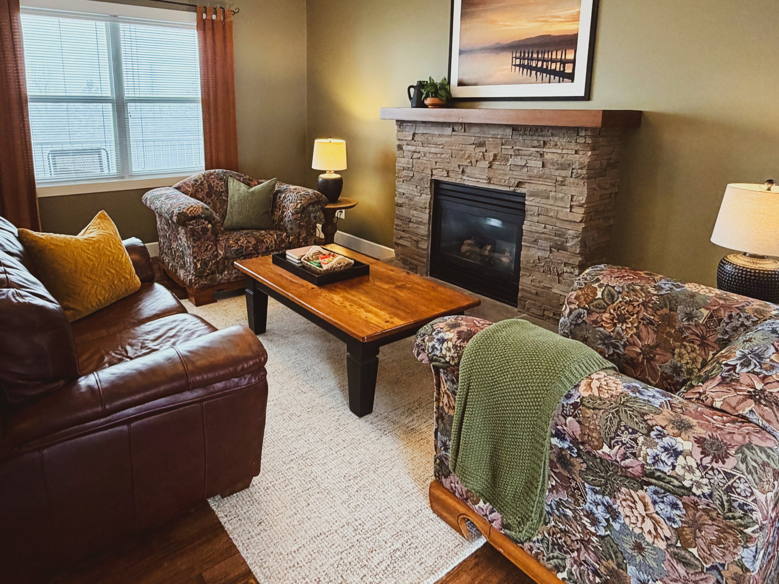 Living room with a window, a stone fireplace with a wooden mantle, floral patterned sofas, a wooden coffee table, lamps, and wall art of a dock on water during sunset.