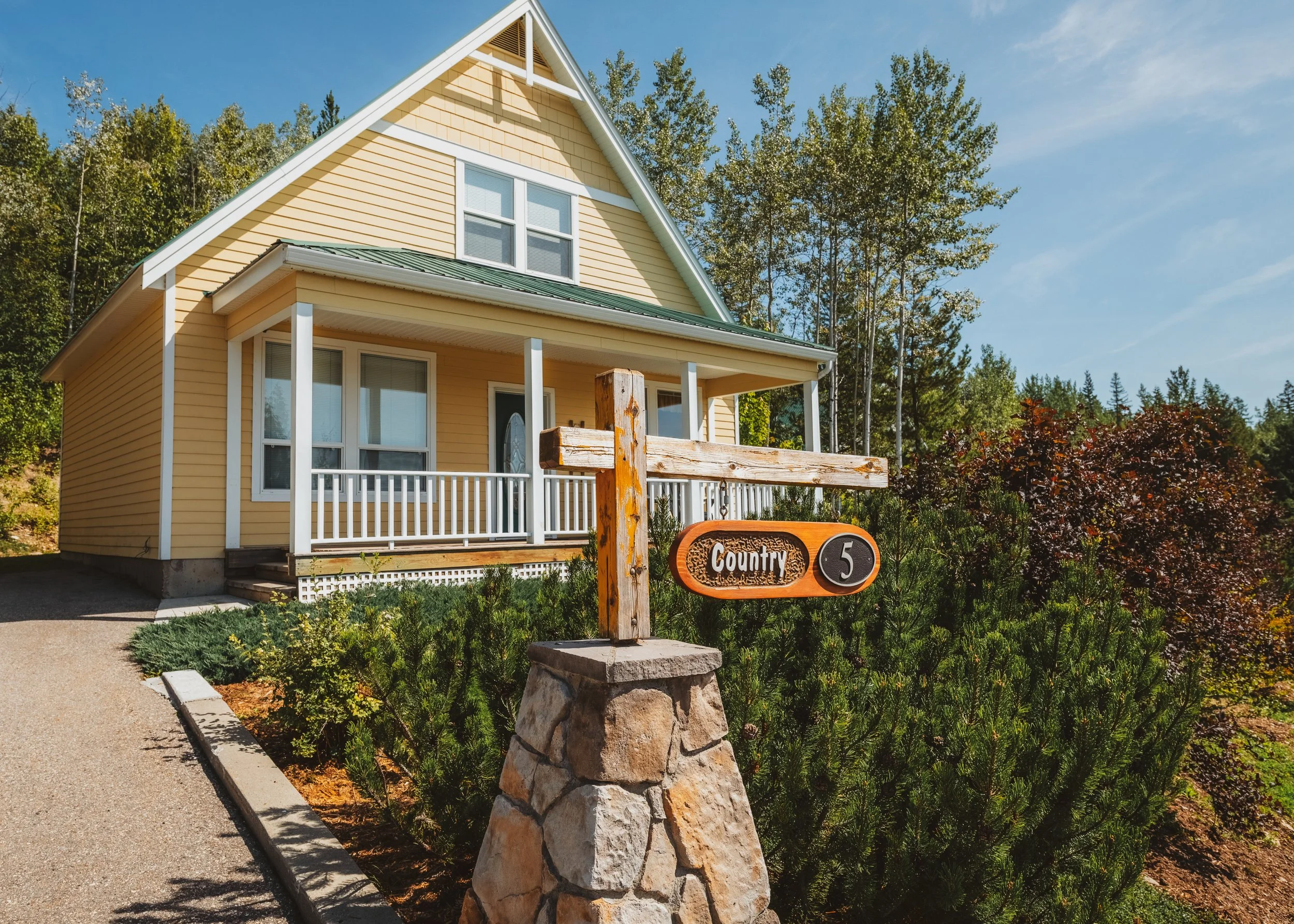 Yellow house with white trim and a green metal roof, surrounded by trees, with a front porch and a wooden sign that reads 'Country 5' on a stone base.