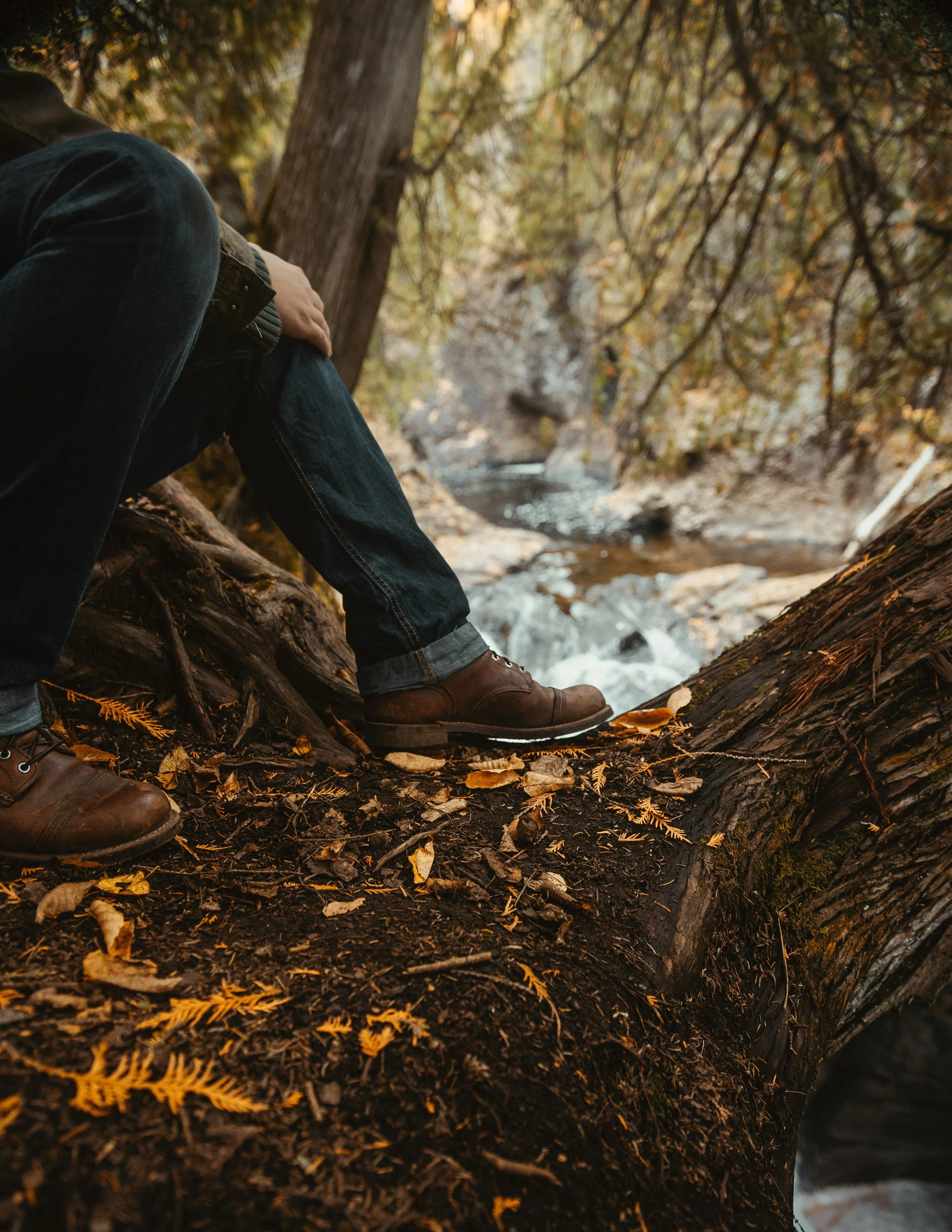 A person sitting on a fallen log by a creek in a forest during autumn, wearing dark jeans and brown boots.