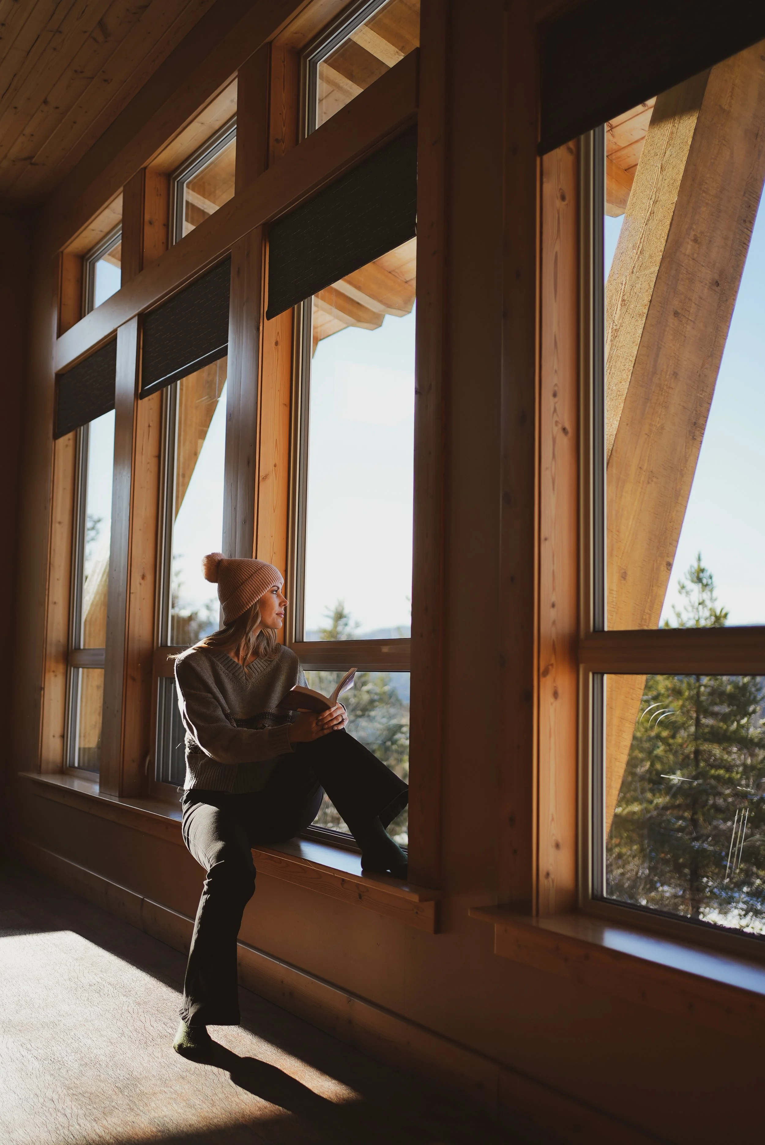 A woman in a beanie and sweater is sitting in front of large windows made of wood and glass, reading a book. Sunlight filters through the windows, illuminating the interior of a wooden building with trees visible outside.