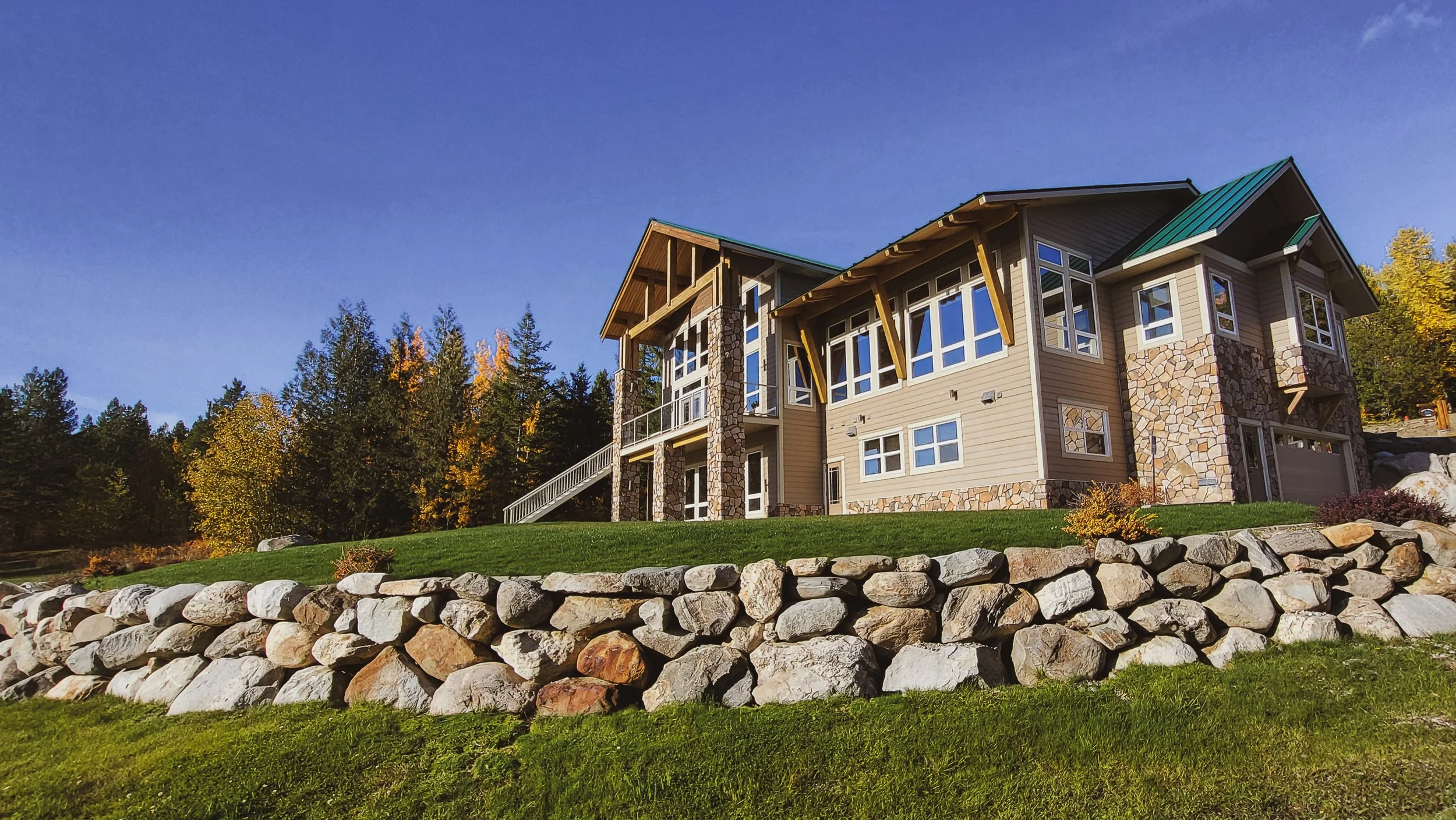 A large modern lodge with multiple windows, stone accents, and a green metal roof, situated on a grassy hill with a stone wall and trees in the background.