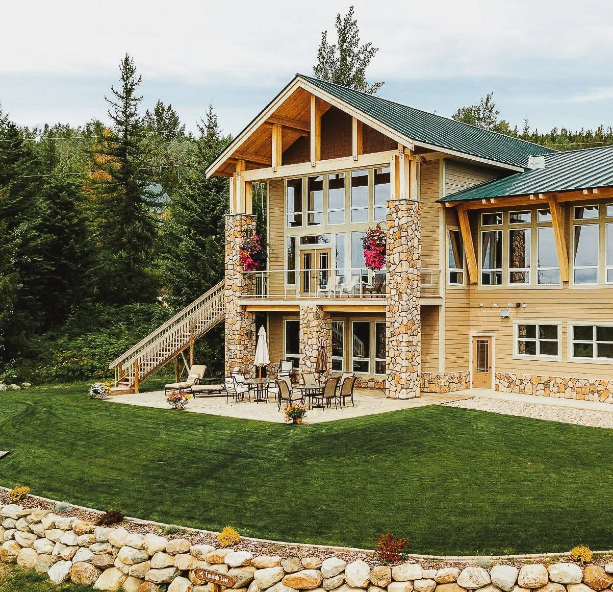 A multi-story modern house with large glass windows, stone pillars, a green metal roof, and a spacious patio with outdoor furniture, surrounded by lush green trees and a well-maintained lawn.