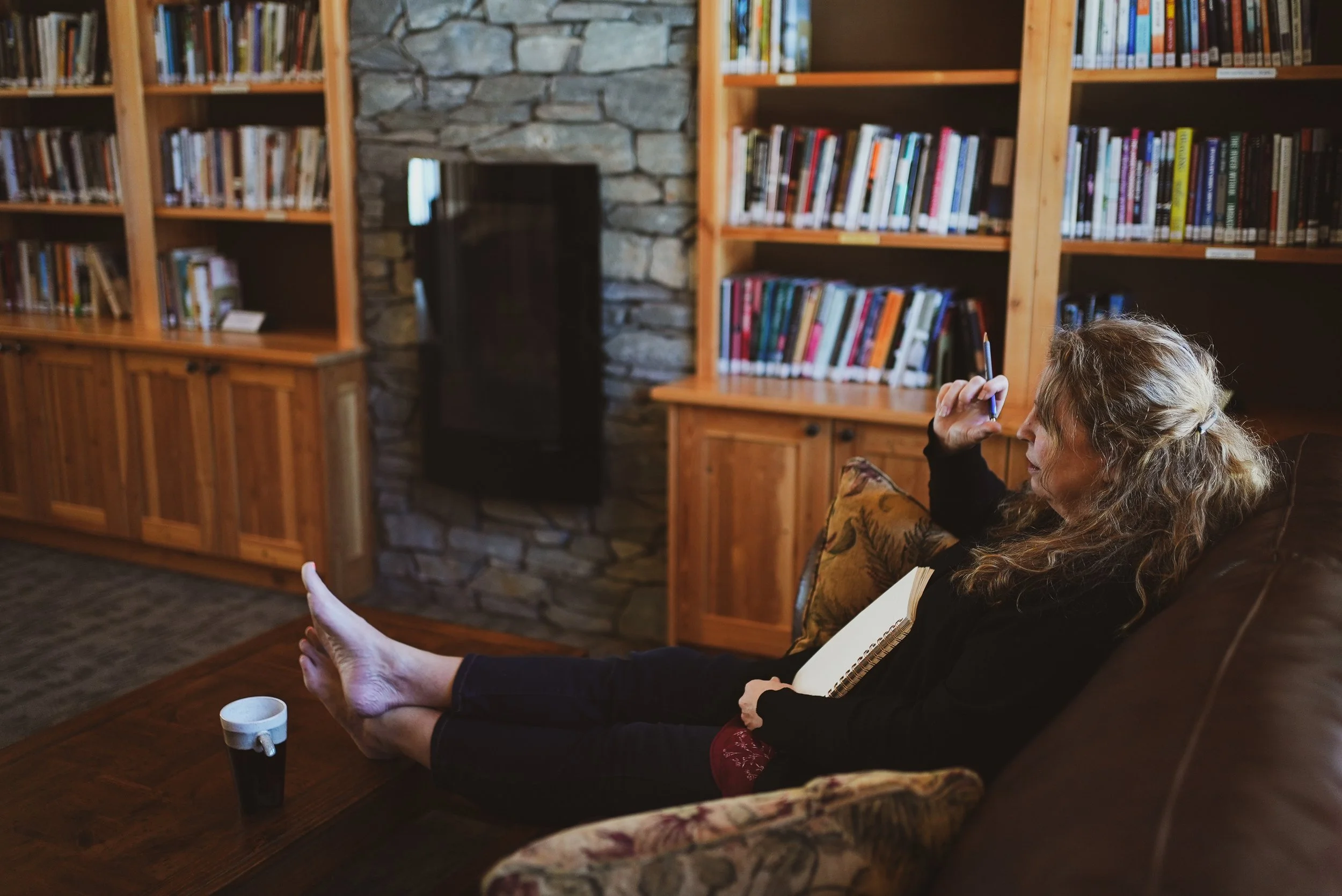 A woman sitting on a sofa with a notebook on her lap, in a cozy library with wooden shelves filled with books and a stone fireplace in the background.