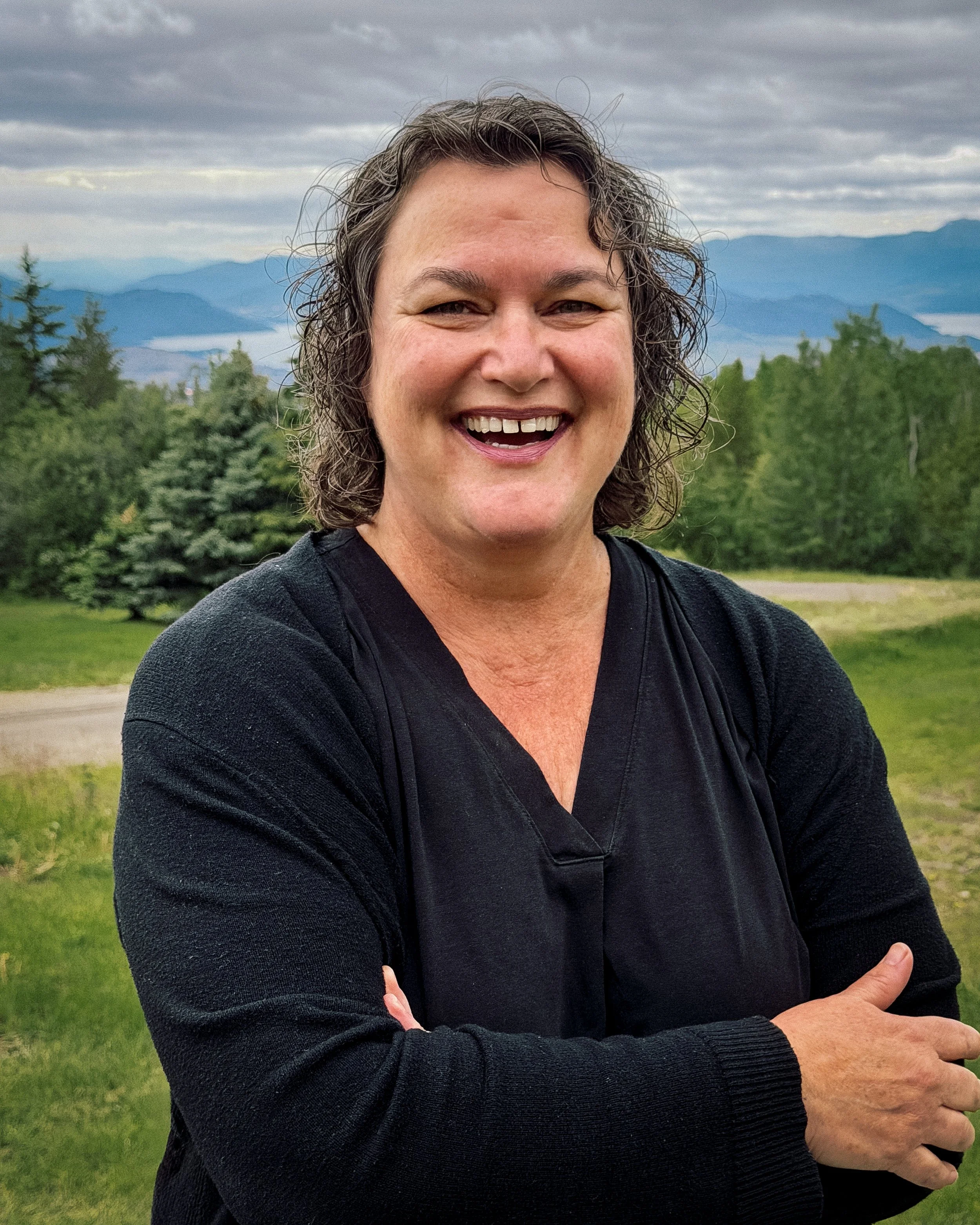 A smiling woman with short dark hair, wearing a black long-sleeve shirt, standing outdoors in front of a scenic landscape with green trees, mountains, and cloudy sky.