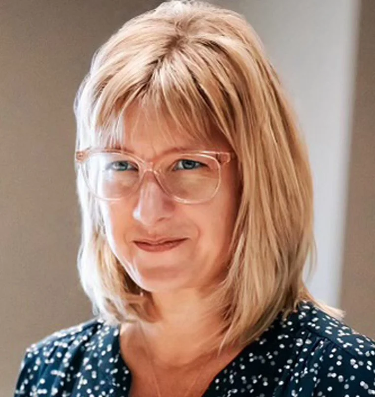 Woman with blonde hair, glasses, wearing a polka dot blouse, smiling indoors.