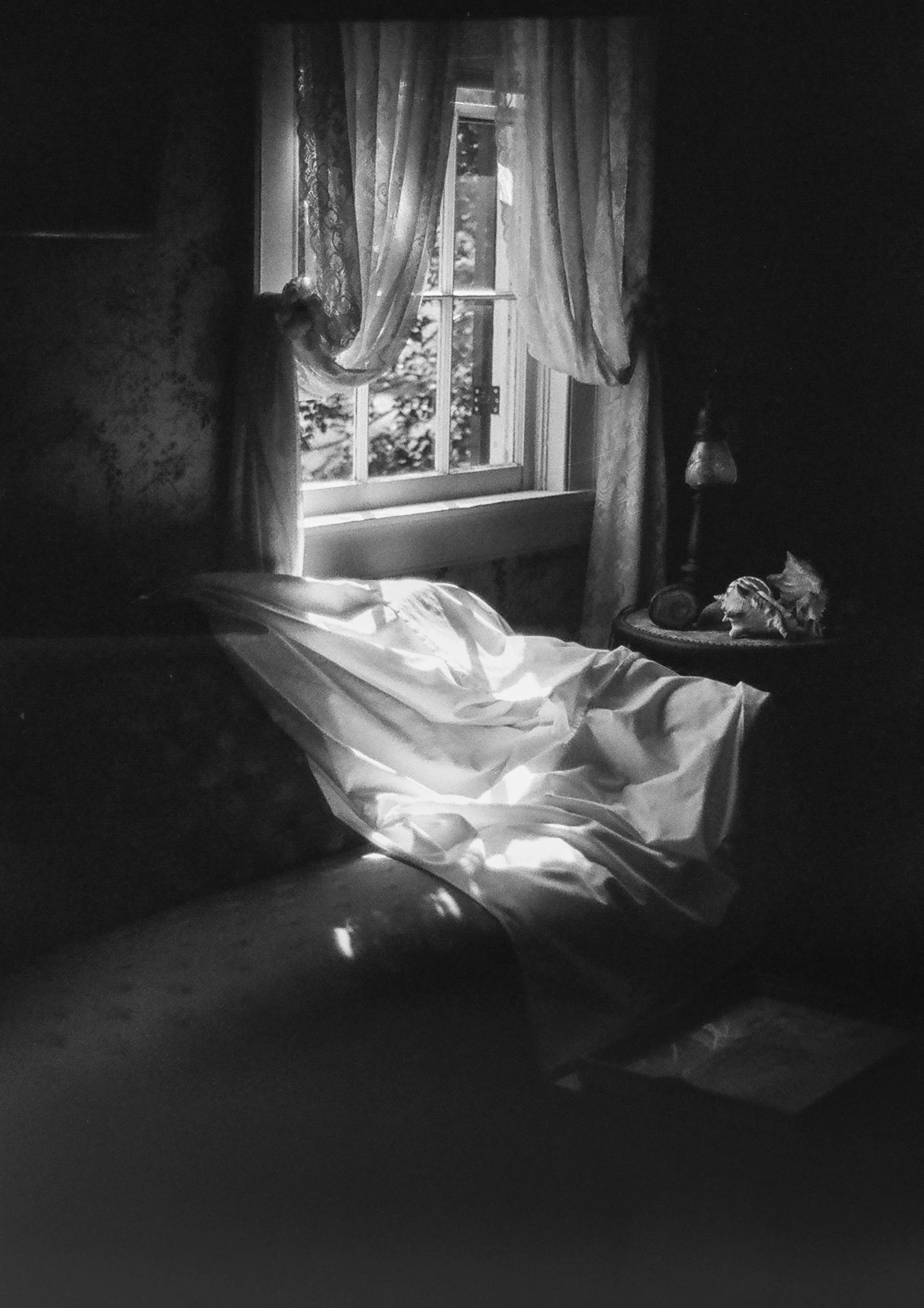 A black-and-white photo of a bedroom with sunlight coming through a window, partially covered by curtains. The bed has a white cover, and there is a lamp and a decorative item on a side table.