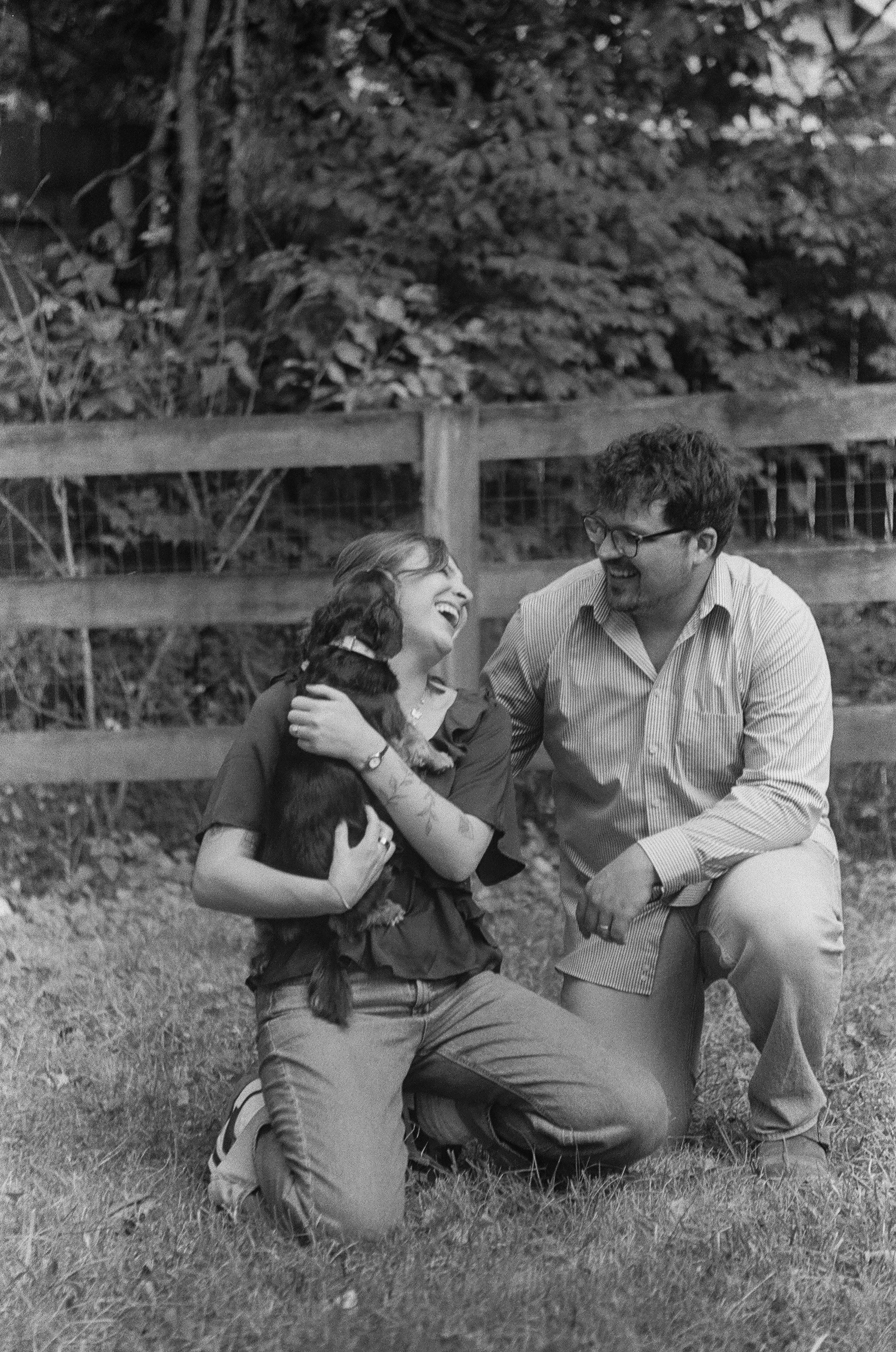 A woman and a man laughing and smiling outdoors, with the woman kneeling on the grass and the man crouching, with a wooden fence and dense trees in the background.