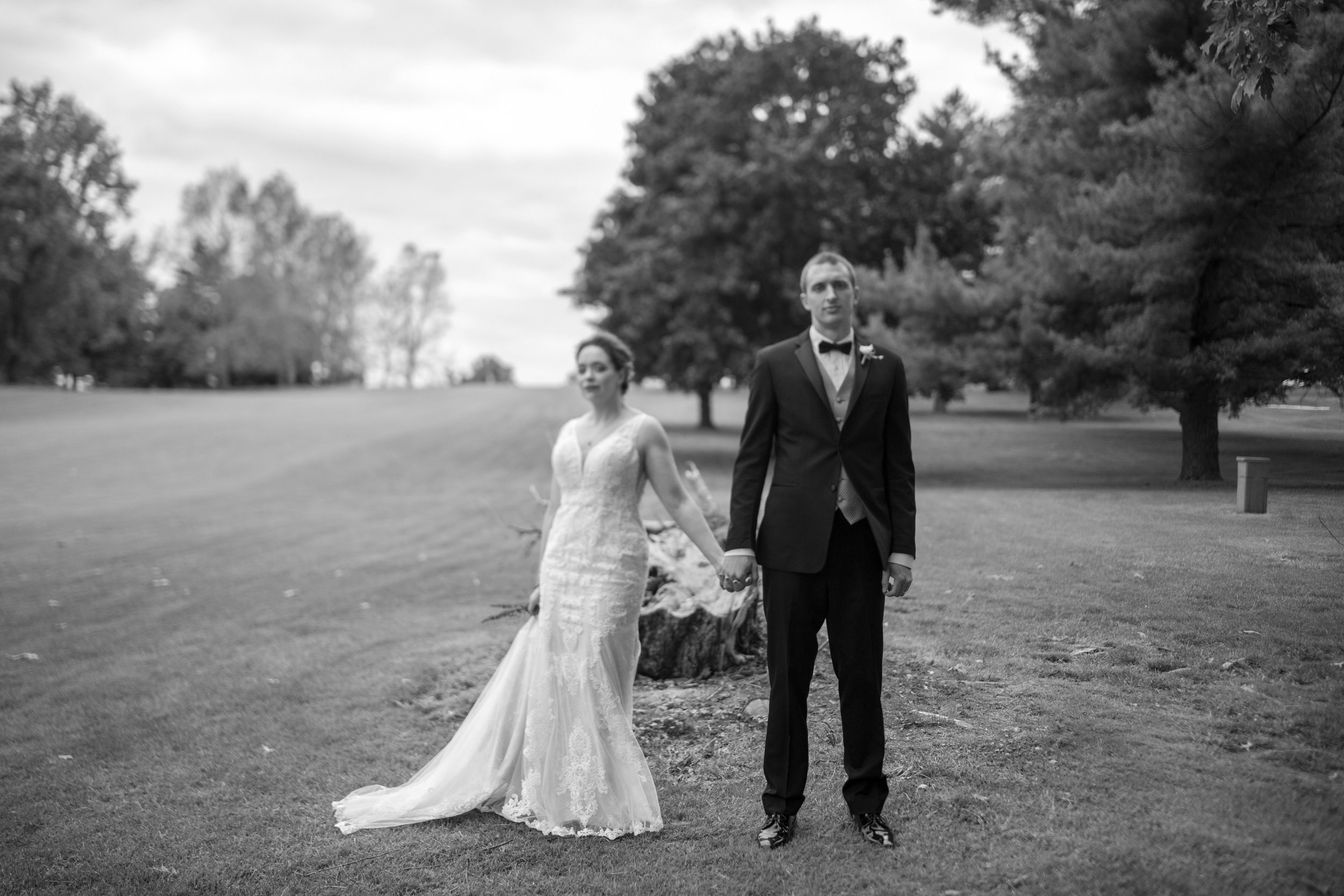 A black-and-white photo of a bride and groom holding hands outdoors in a park with trees in the background. The bride is wearing a wedding dress and the groom is dressed in a tuxedo with a bow tie.