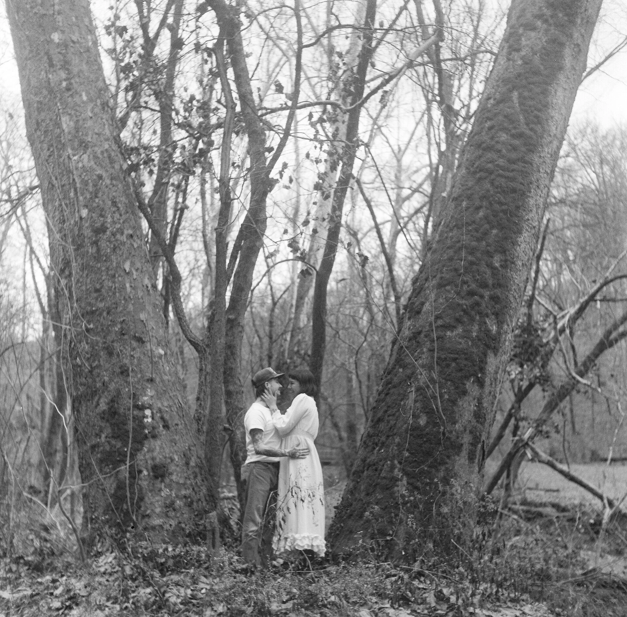 A black and white photo of a couple dressed in wedding attire standing closely, holding hands, and sharing a kiss in a wooded outdoor area with trees and fallen leaves.