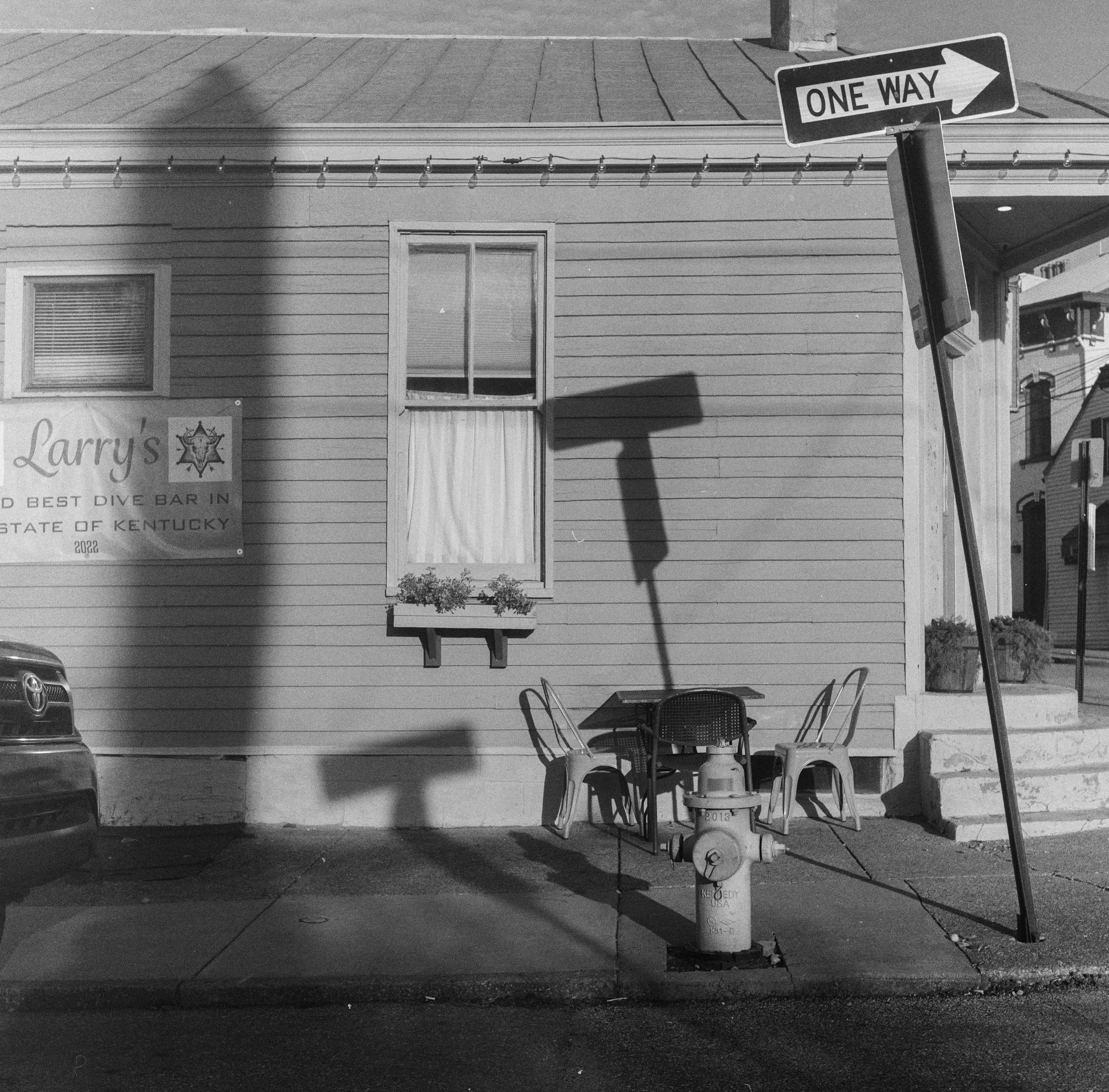 A black-and-white photo of the side of a building with two windows, a string of holiday lights, and a sign that reads 'One Way' with an arrow pointing to the right. There is a sidewalk and some outdoor furniture, including chairs and a table, in the 